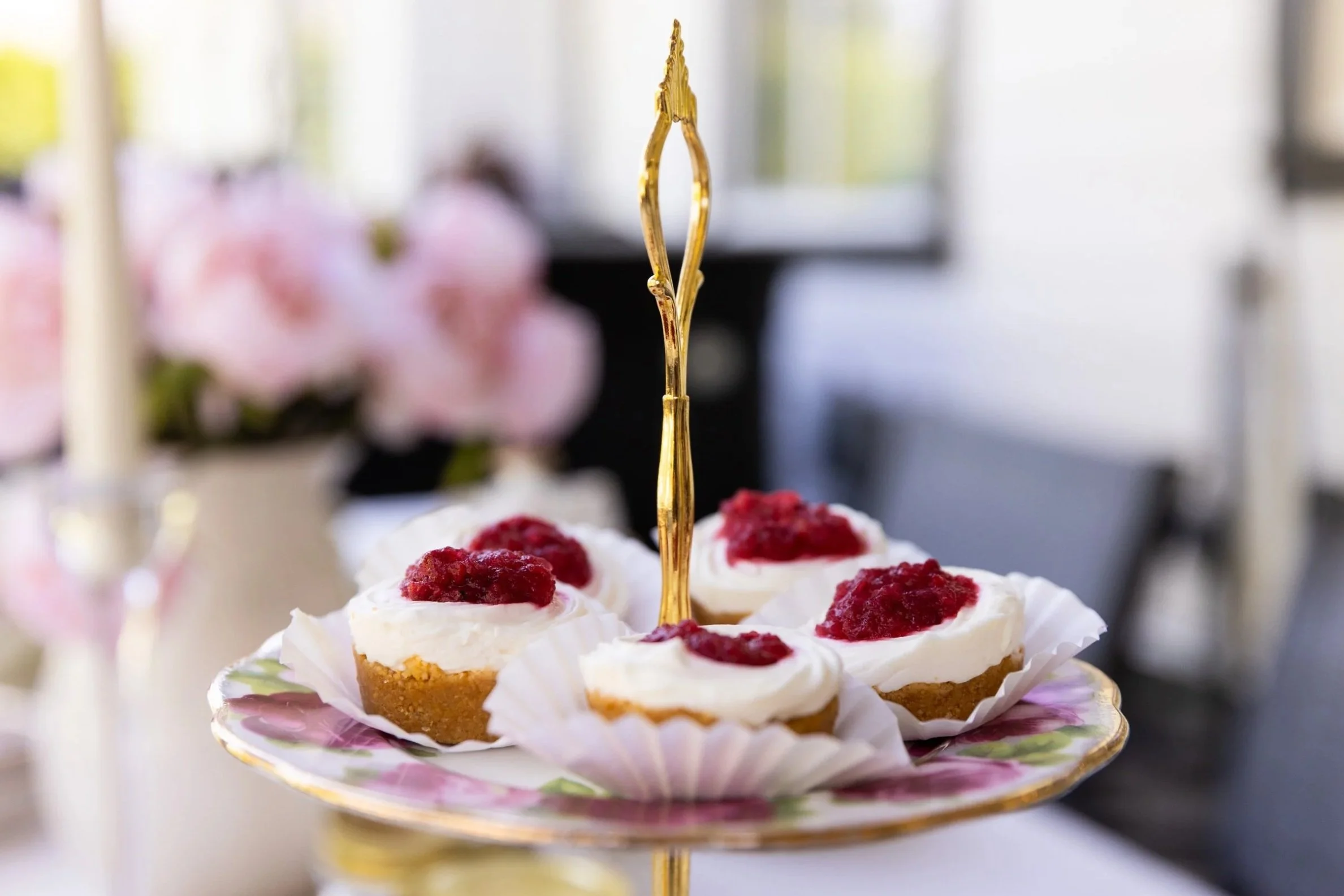Close-up of a serving plate with mini cheesecakes topped with raspberry jam, topped with a decorative gold fork, blurred background of a room with pink flowers and natural light.