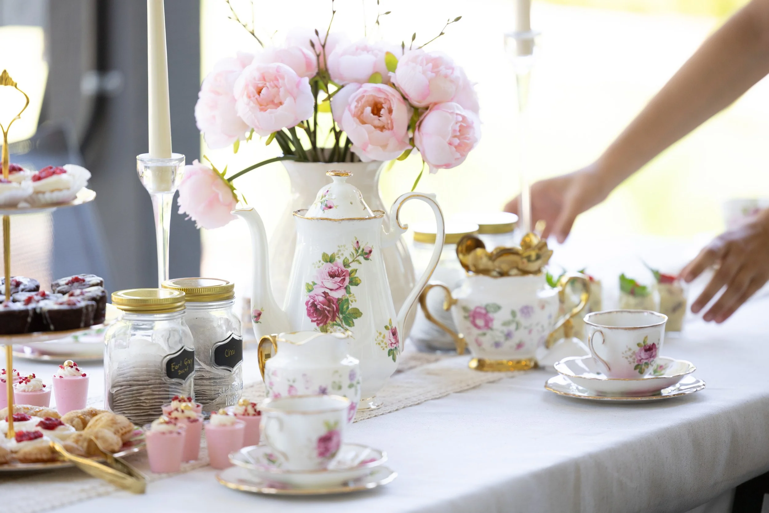A table set for tea with pink peonies in a floral teapot, and various desserts including cupcakes, cookies, and jam jars, in a bright environment with sunlight.