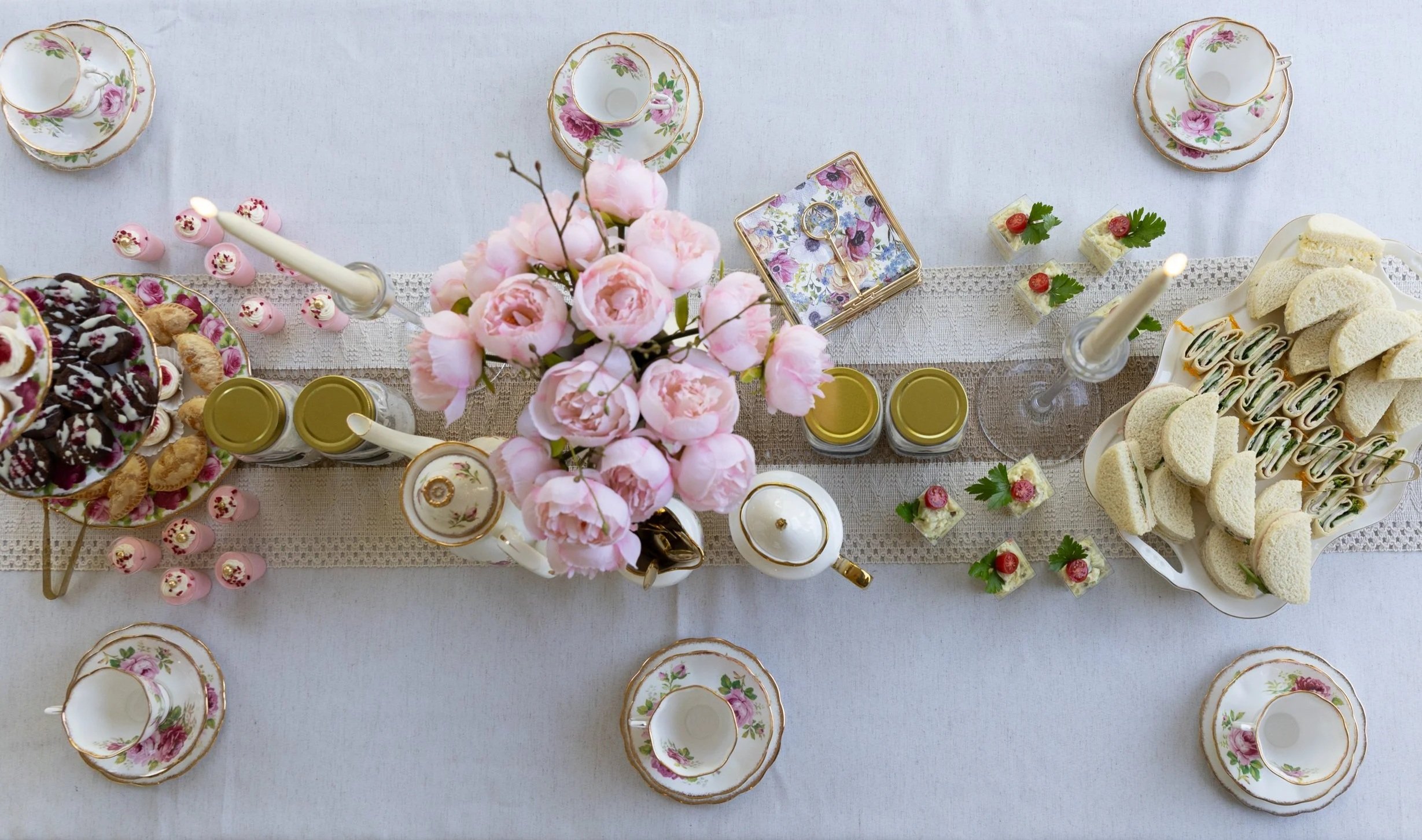 An elegant table set for a tea party with floral china dishes, pink roses as a centerpiece, and an array of sweet and savory finger foods including sandwiches, pastries, and small desserts.