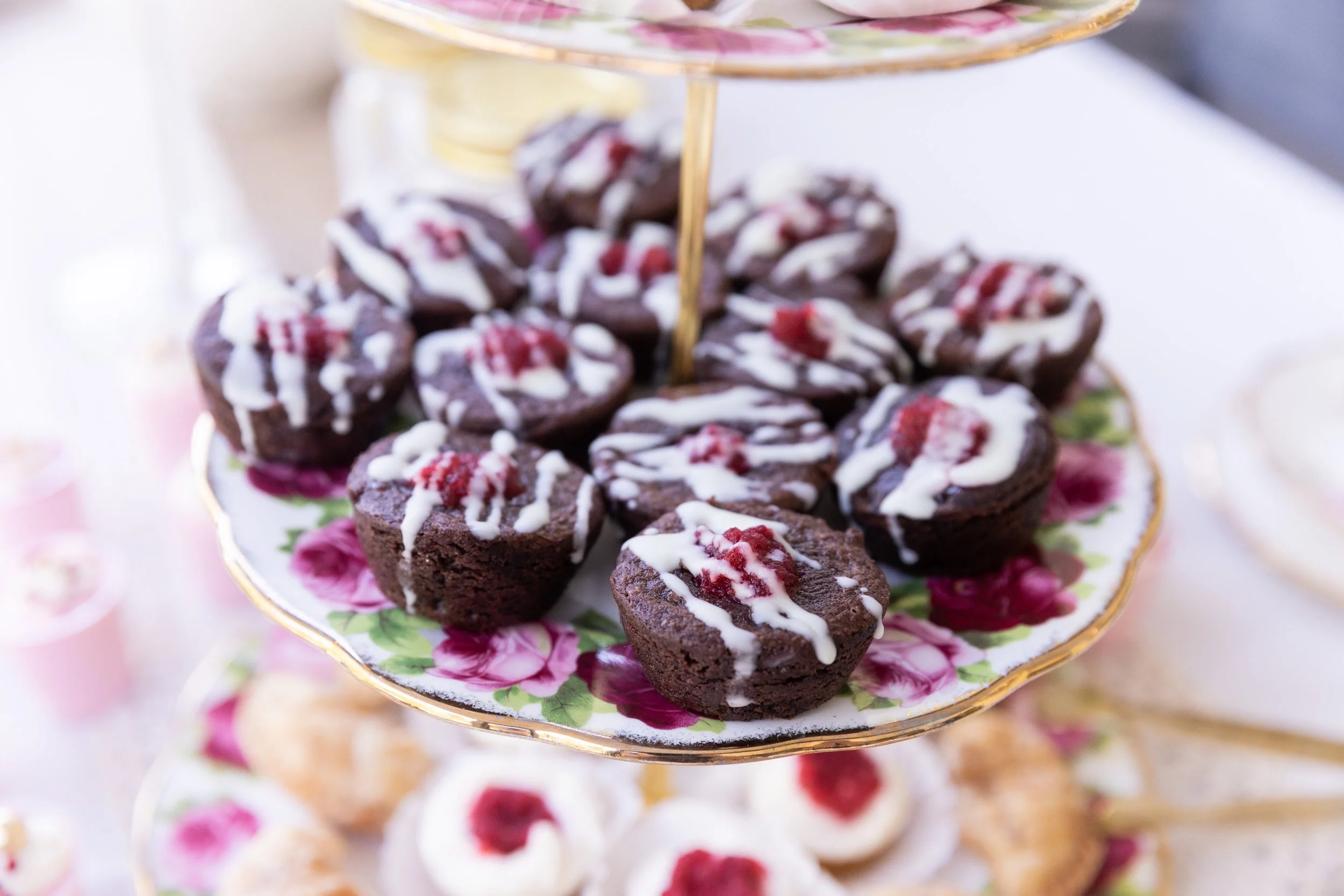 Close-up of a three-tiered decorative serving stand filled with assorted chocolate desserts topped with white drizzle and red fruit, on a floral-patterned plate.