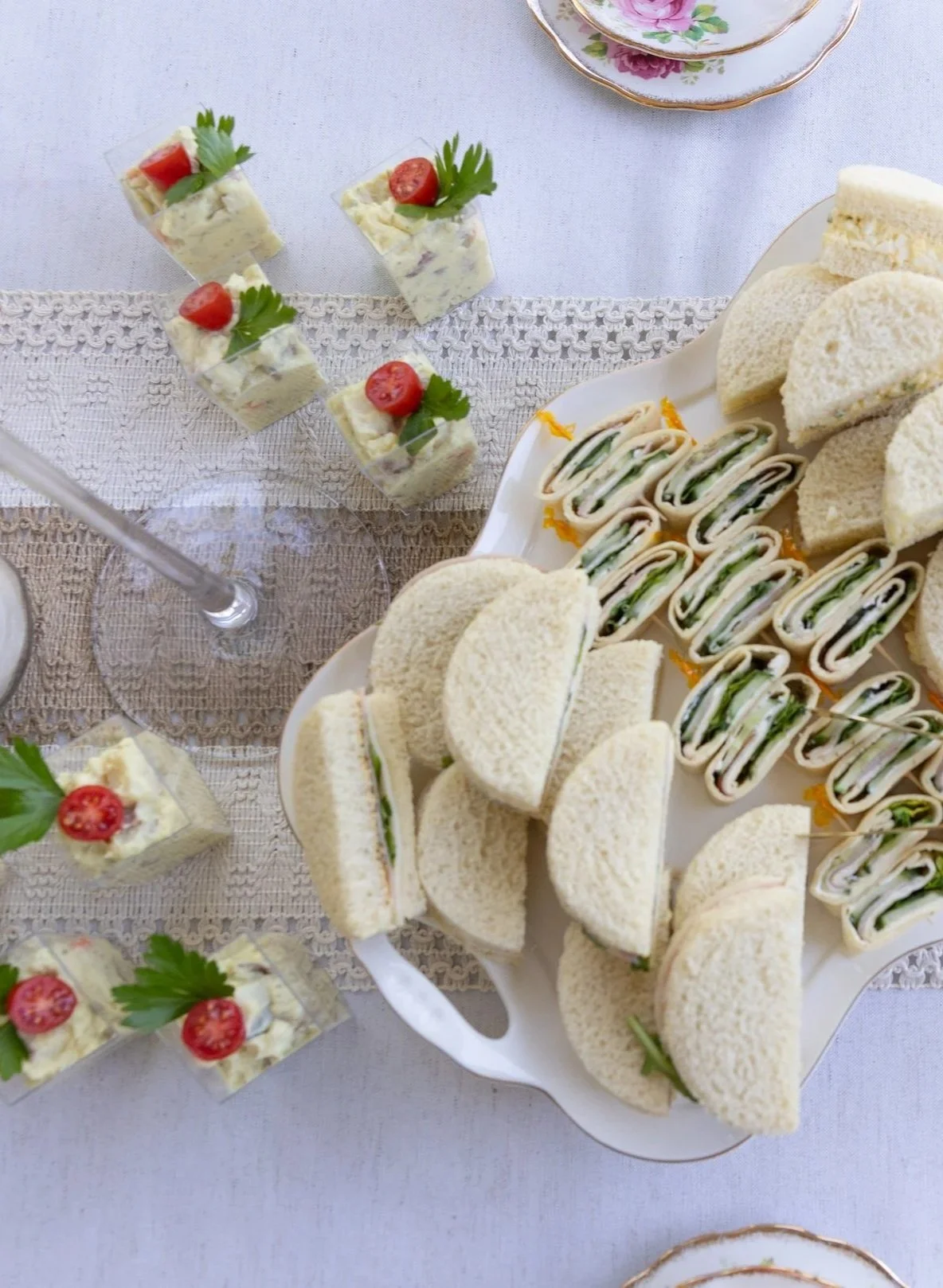 Spread of tea sandwiches on a white serving tray and small layered appetizer squares topped with cherry tomatoes and parsley on a lace table runner.