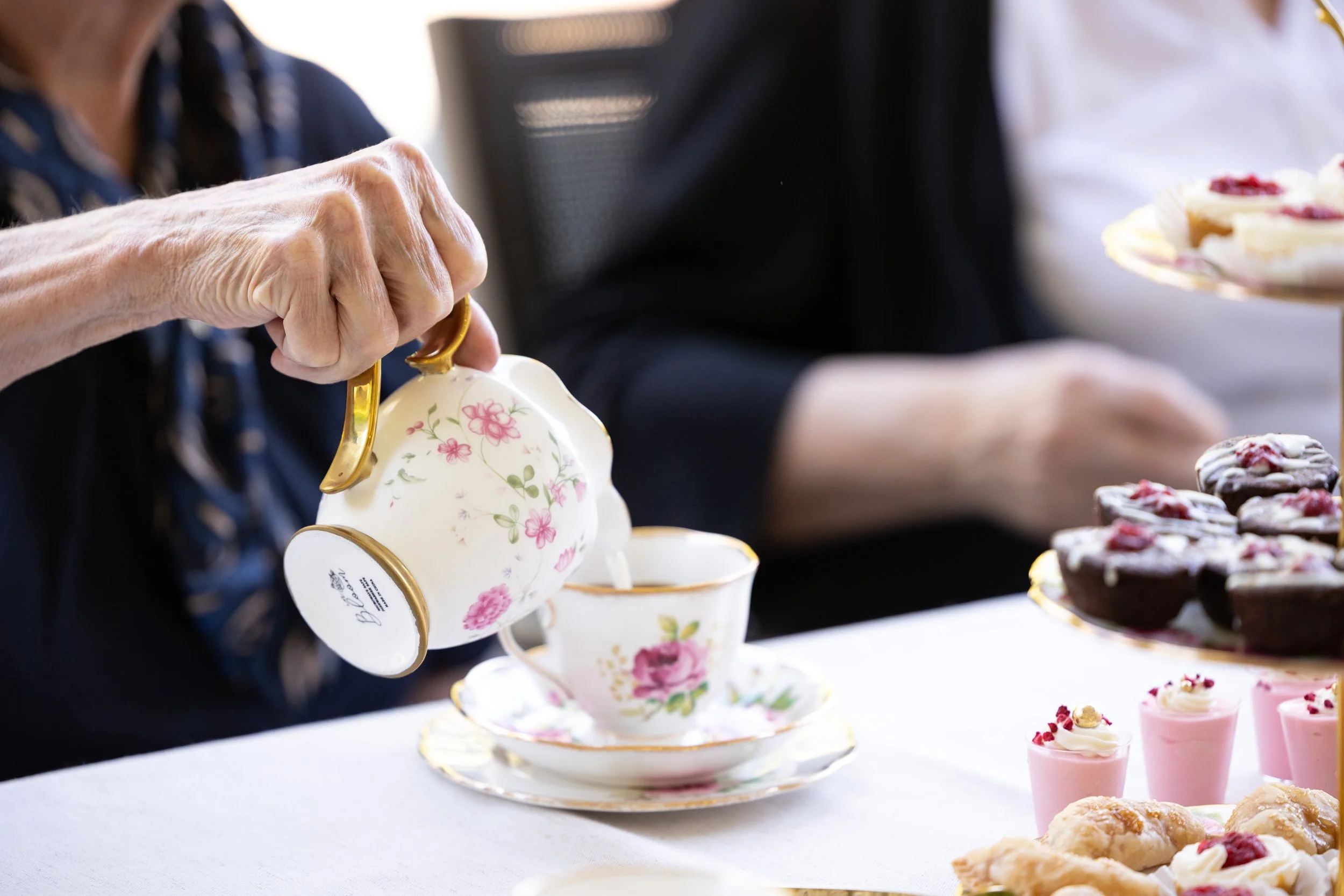 A person's hand pouring tea from a floral teapot into a cup, with a table of cupcakes and desserts in the background.