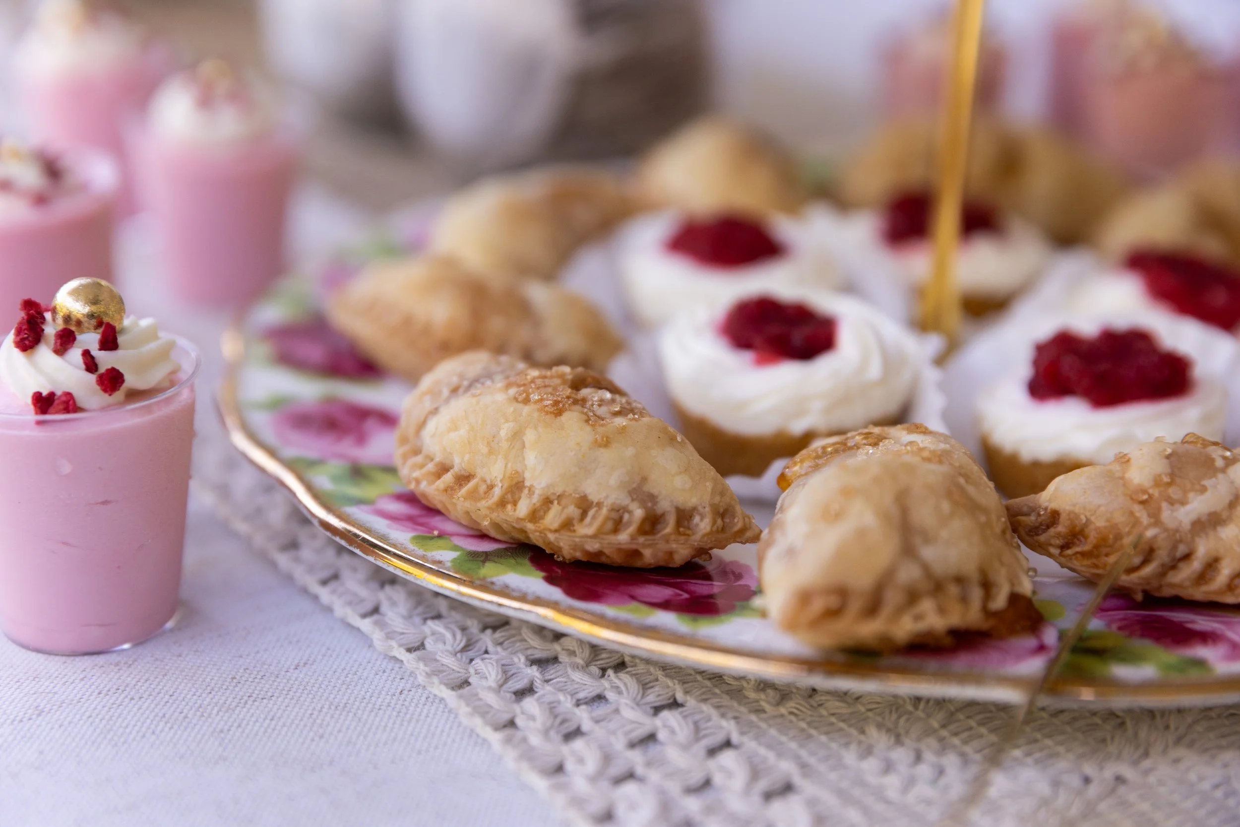 Close-up of pink dessert glasses topped with whipped cream, red sprinkles, and a gold edible sphere, set on a decorative plate with assorted pastries including cream puffs with raspberry topping, on a lace tablecloth.