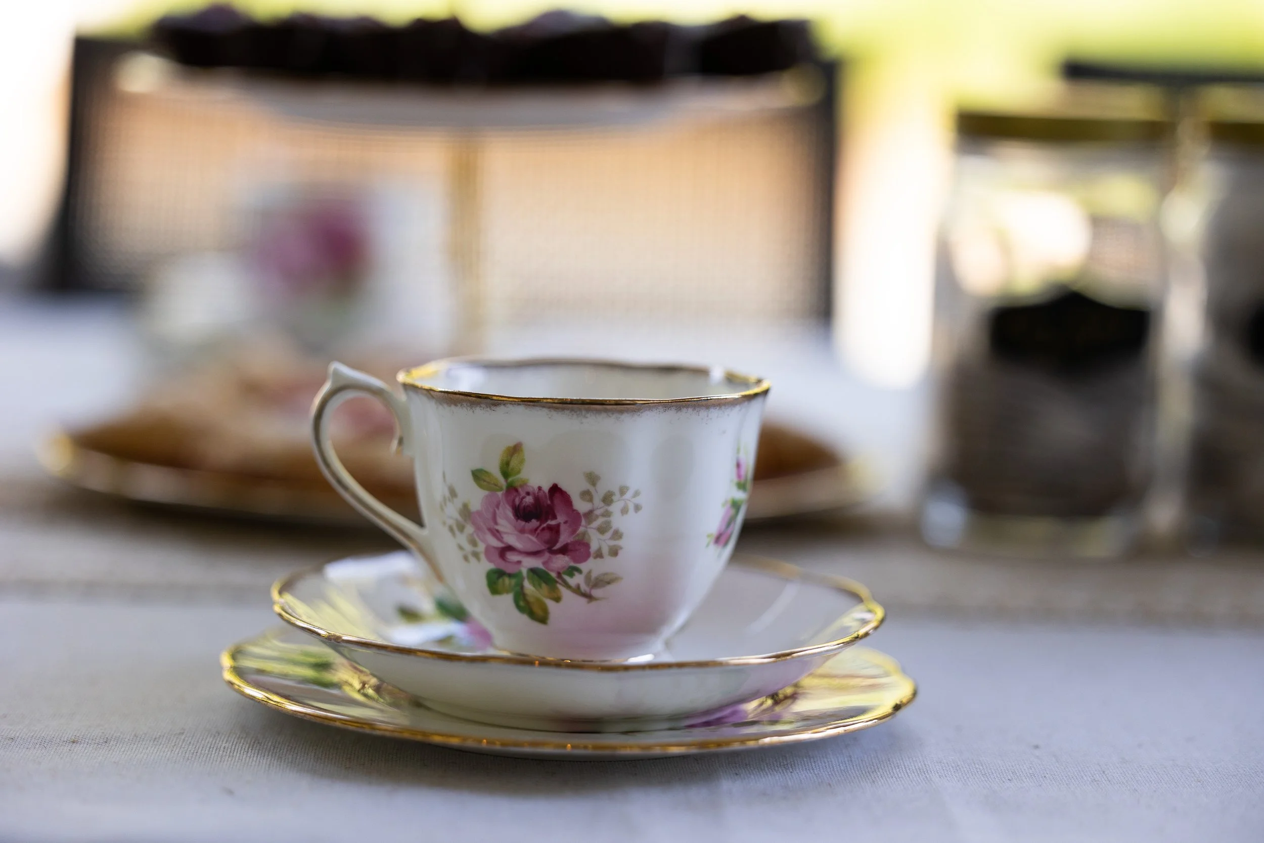 A close-up of a vintage floral teacup with a matching saucer on a table, with a blurred background featuring other dishes and kitchen items.