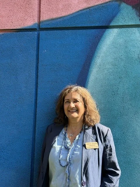 Woman with curly hair smiling, standing in front of a colorful mural with blue and pink shapes