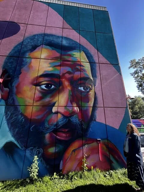 Colorful mural of a man's face with a contemplative expression, painted on the side of a building, with a woman standing nearby.