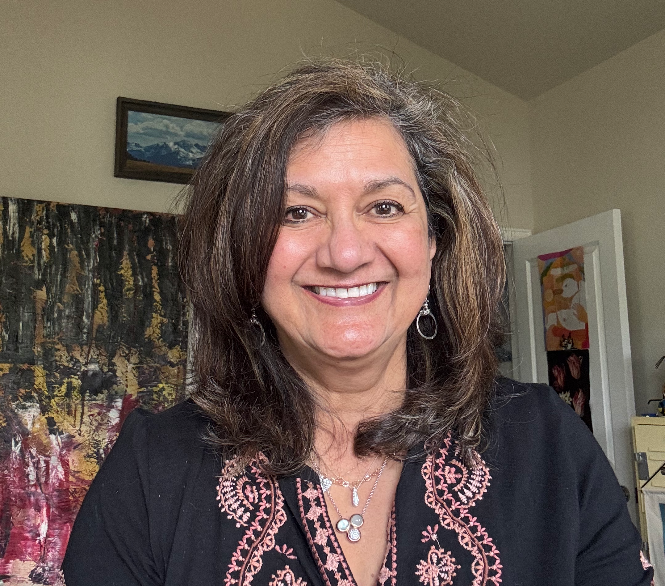 Close-up of a smiling woman with brown curly hair wearing a black top with intricate pink embroidery and jewelry, standing indoors with artwork and furniture in the background.