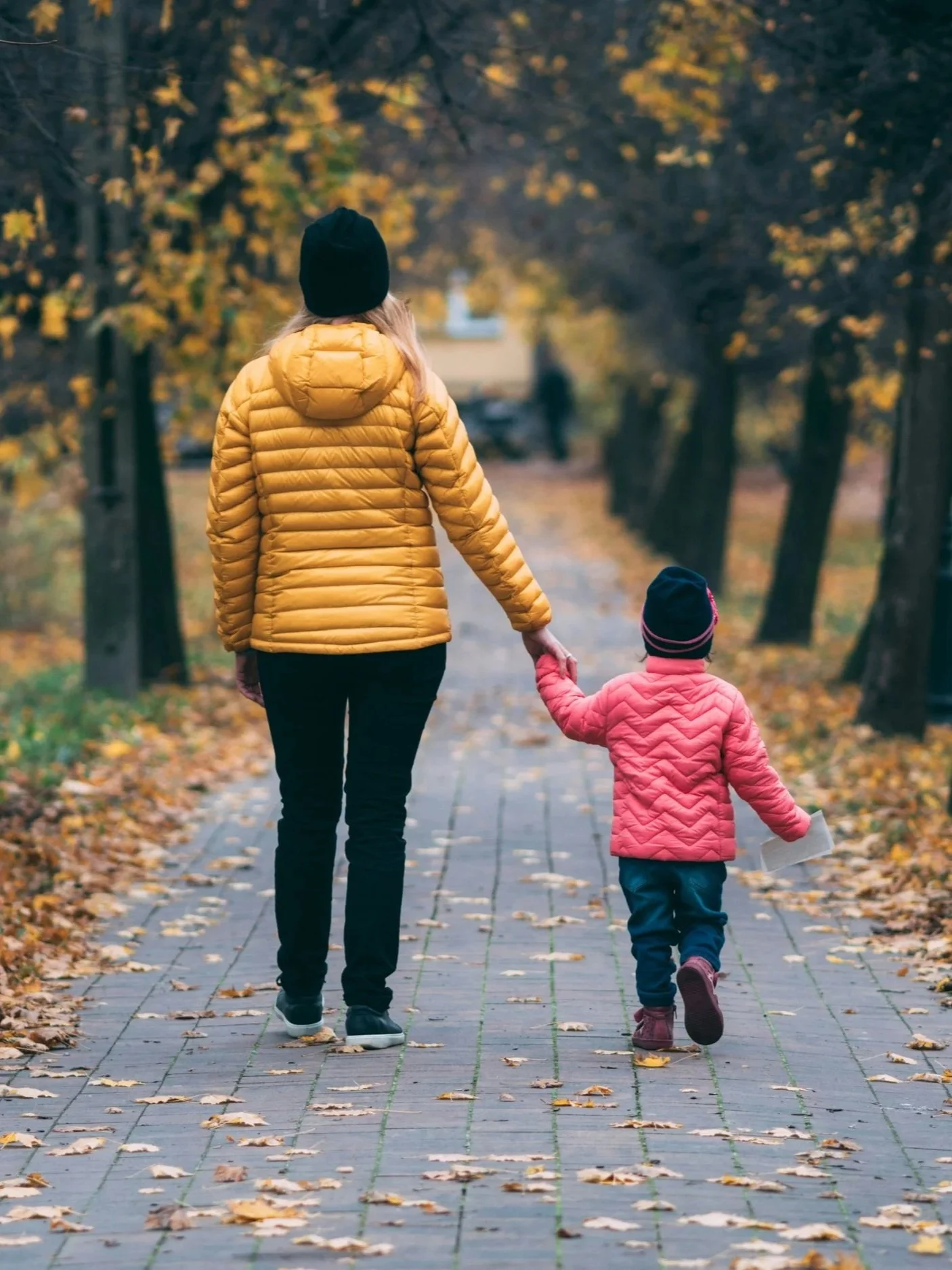 A woman and a child walking hand in hand on a leaf-covered sidewalk in autumn, wearing bright jackets and beanies.