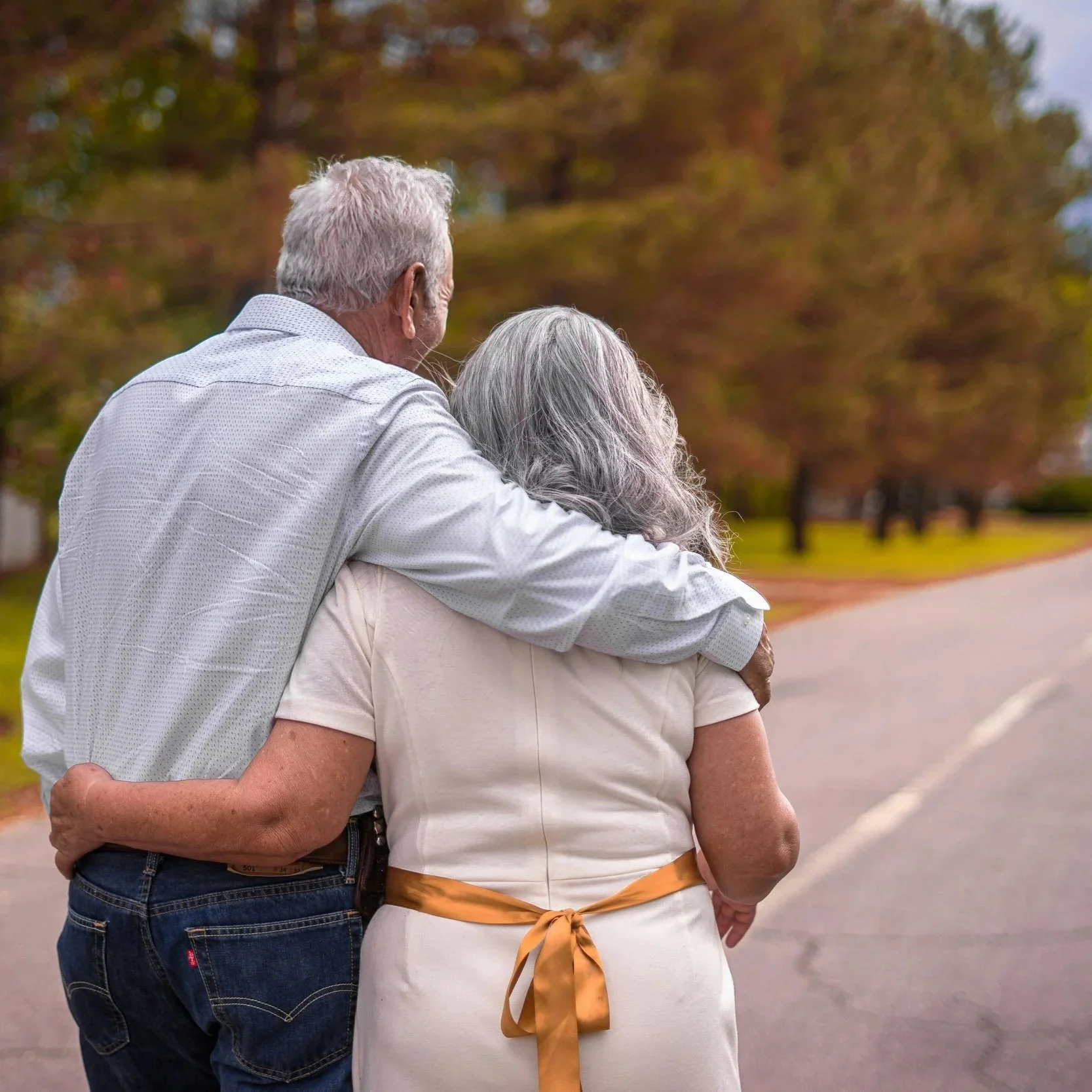 An elderly man and woman hugging outdoors with autumn trees in the background.