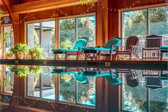 Indoor swimming pool area with wooden walls, large windows, and chairs along the poolside. Plants are on a table near the windows, and the pool water reflects the surroundings.