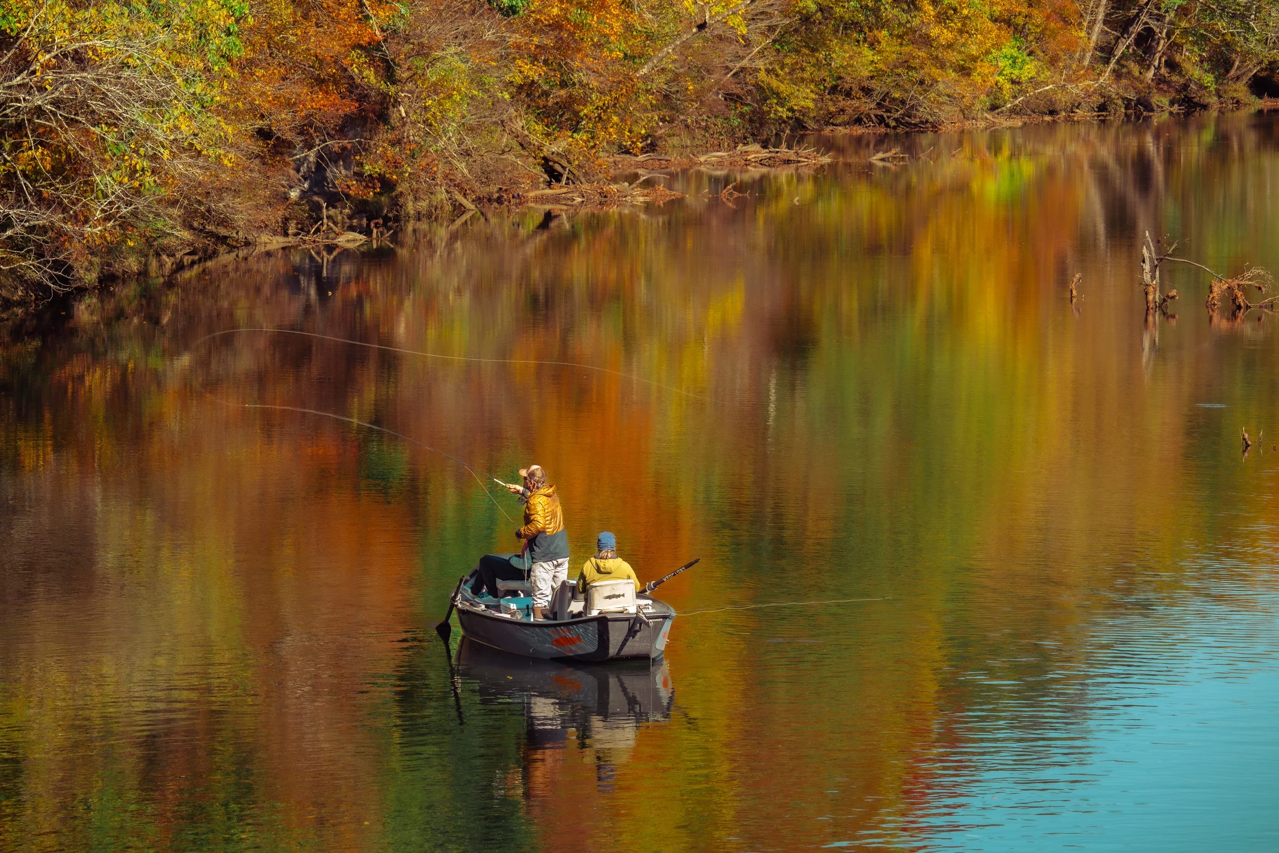 Three people in a small boat on a calm river, surrounded by trees with fall foliage in shades of orange, yellow, and green.