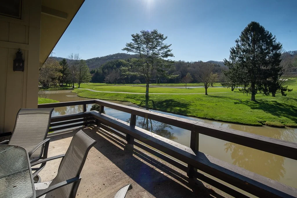 View of a golf course with lush green grass, trees, and a small waterway from a balcony with outdoor chairs and a glass table on a sunny day.
