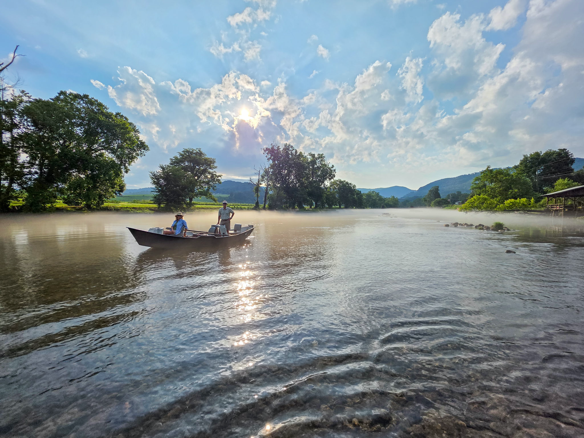 A boat with two people on a river under a partly cloudy sky with the sun shining through the clouds, trees lining the riverbank, and mountains in the background.