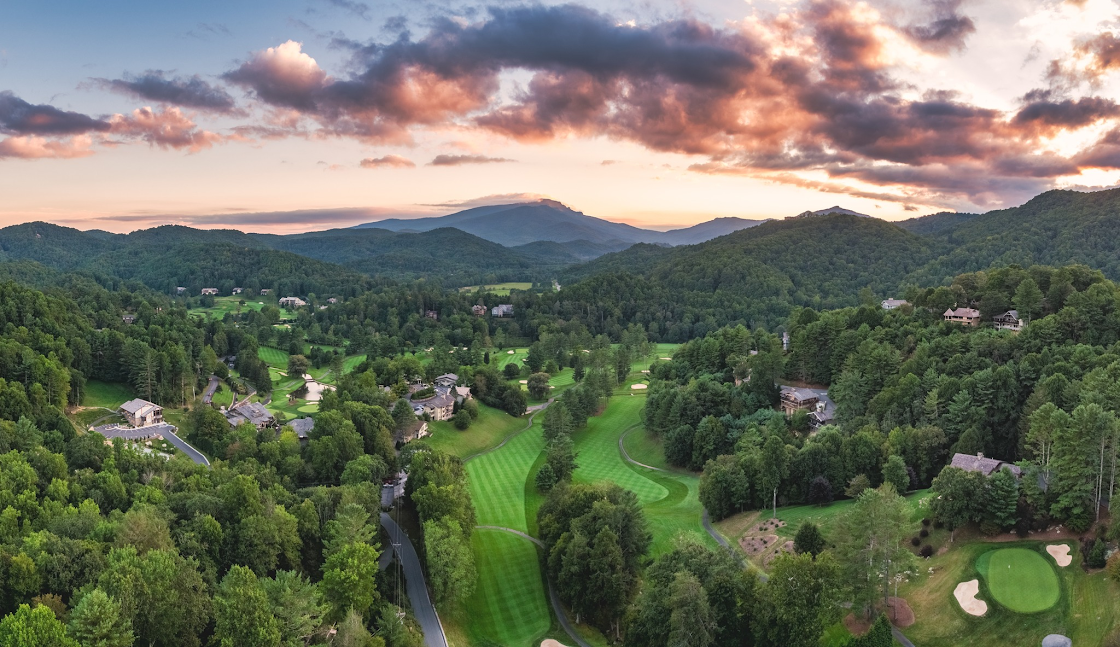 Aerial view of a golf course surrounded by lush green trees and residential houses, with mountains in the background and a colorful sunset sky with clouds.