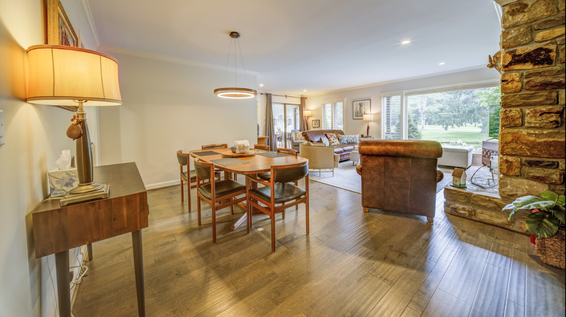 Living room with wood flooring, seating area with sofa, armchair, and coffee table, large windows with views of greenery outside, stone fireplace, and dining area with wooden table and chairs, table lamps and decorative lighting.