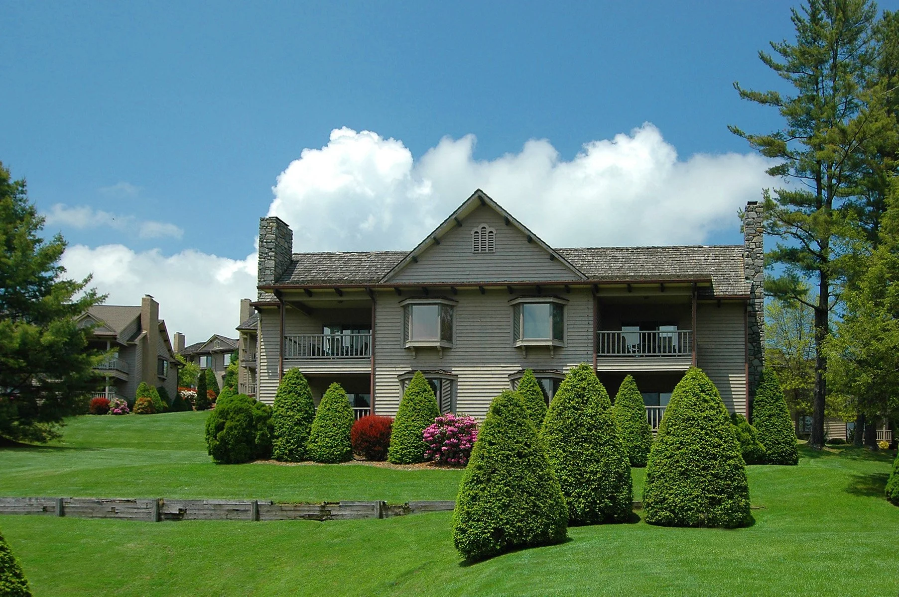 A two-story house with gray siding, front porch, and balconies, surrounded by well-manicured lawn and neatly trimmed bushes, with trees and a blue sky with clouds in the background.