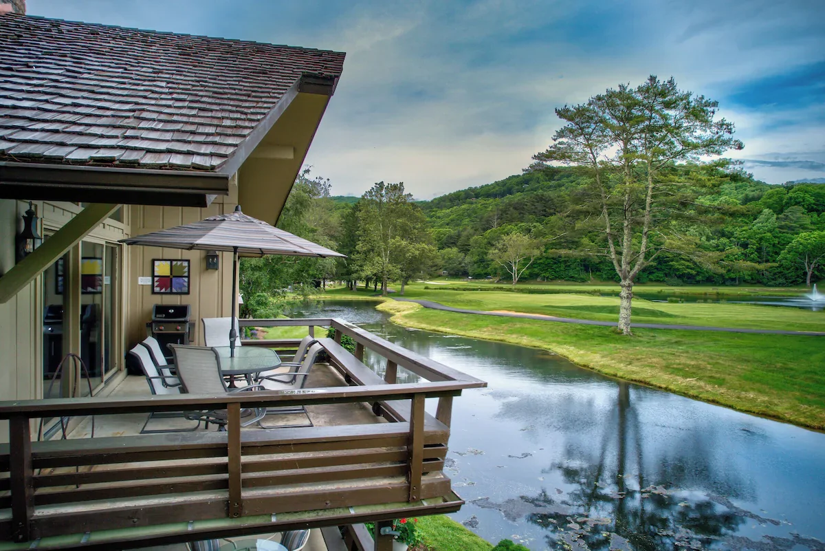 A balcony with a table, chairs, and umbrella overlooking a river and a golf course with trees and hills in the background.