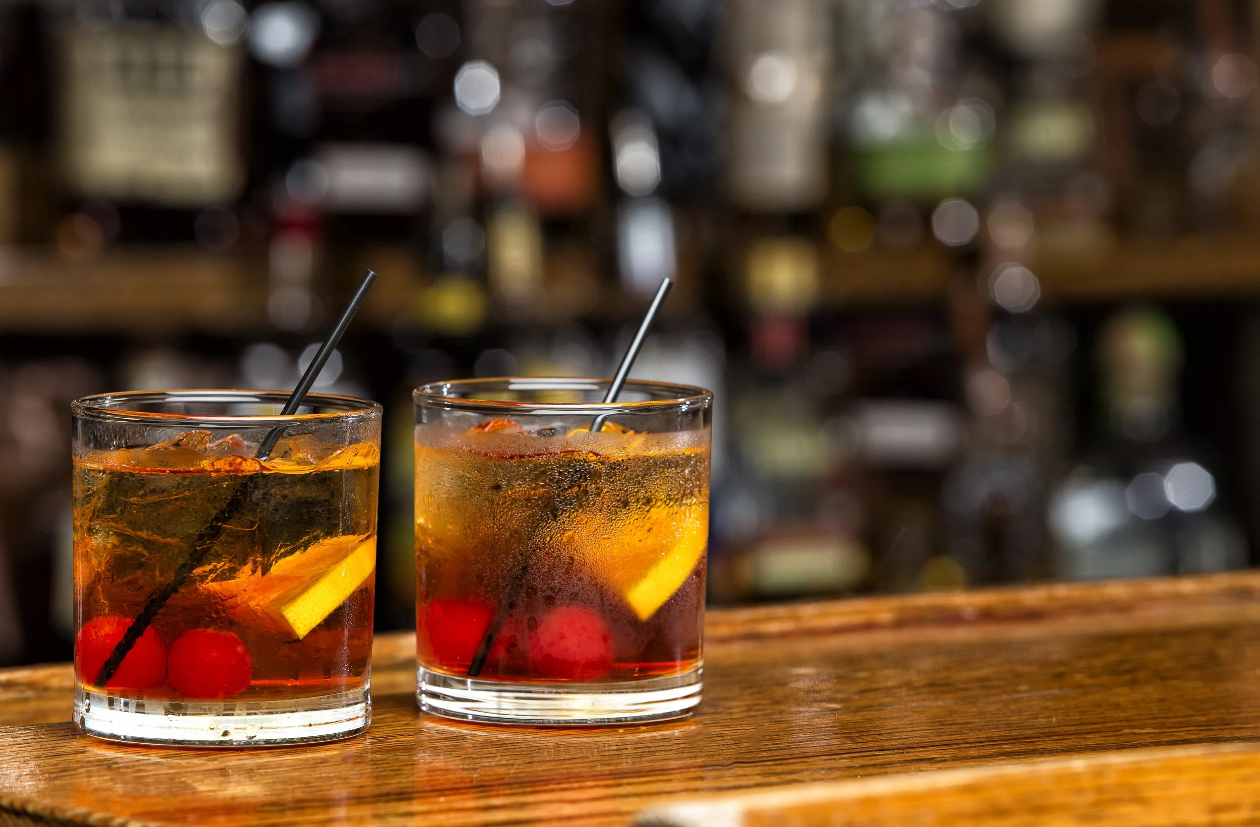 Two glasses of cocktail with ice, lemon slices, cherries, and straws on a wooden bar counter, with a blurred bar background.