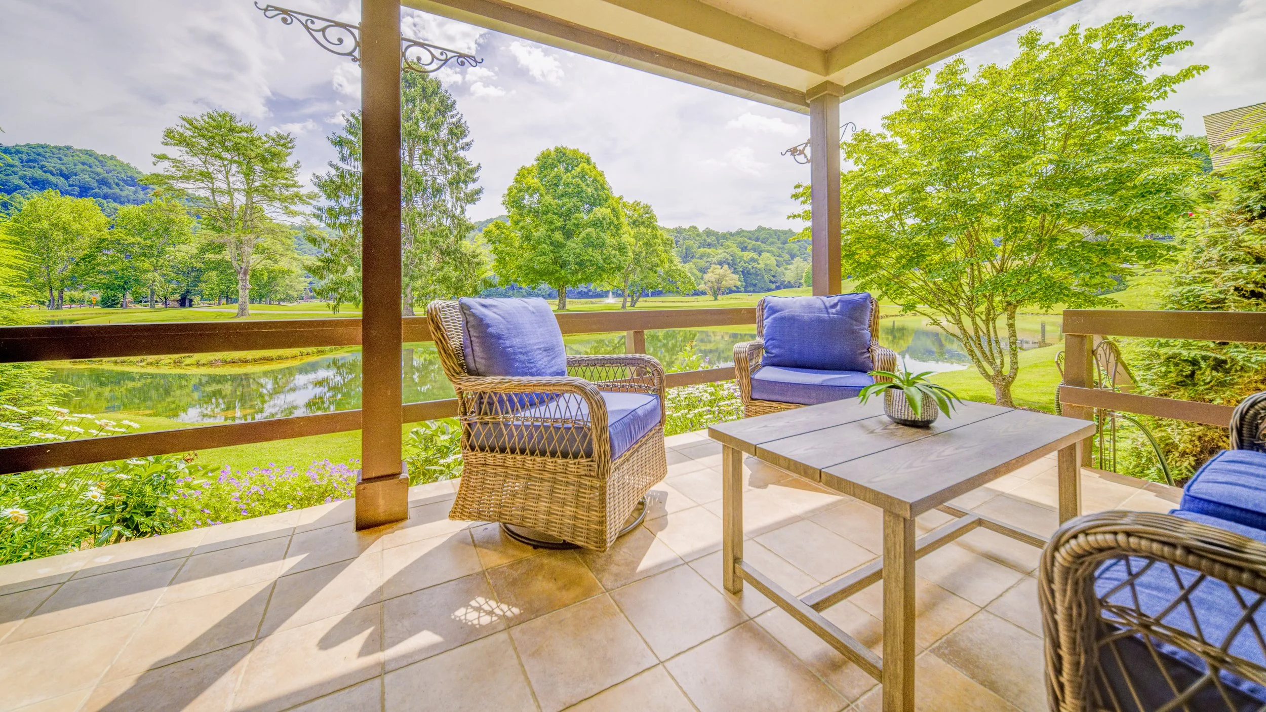 A porch with wicker chairs with blue cushions, a wooden table with a potted plant, overlooking a lake, lush green trees, and distant hills under a partly cloudy sky.