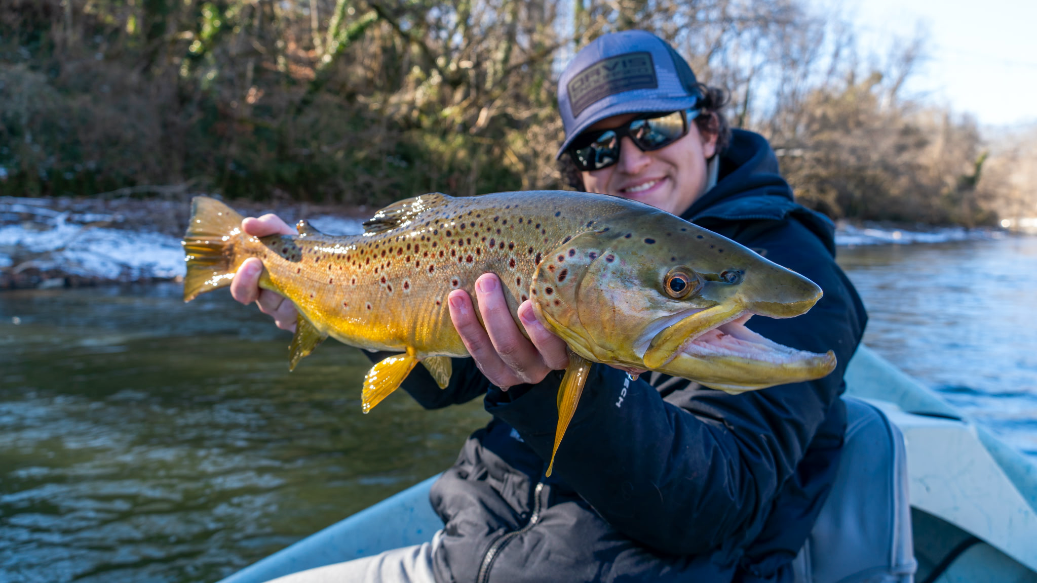 A person wearing a cap, sunglasses, and a jacket holding a large fish with yellow and brown spots in a river with a forested background.