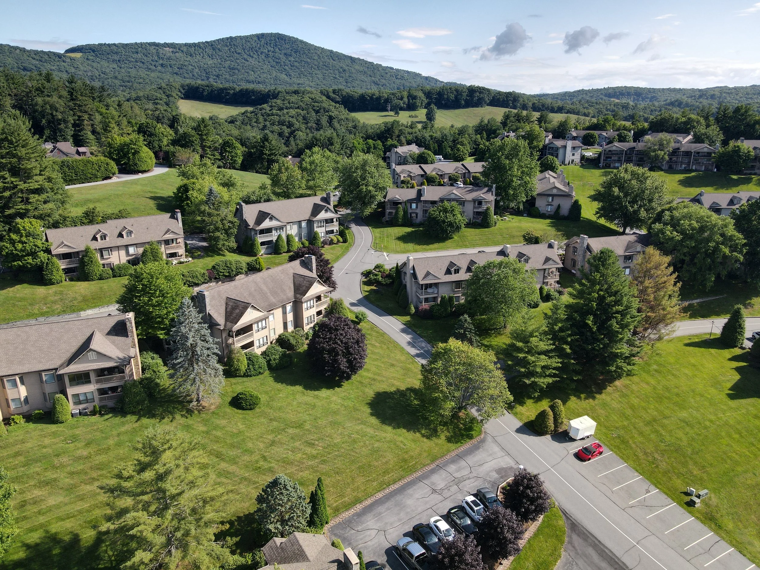 Aerial view of a residential area with multiple apartment buildings surrounded by lush green trees, with mountains in the background and parking lot with cars in the foreground.
