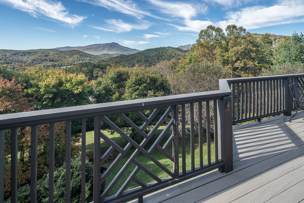 A wooden balcony railing overlooking a lush green forest with mountains in the background under a partly cloudy blue sky.