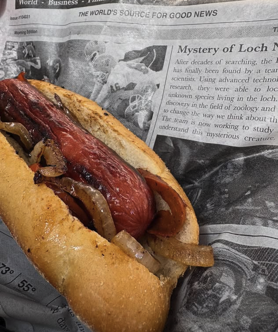 A hot dog with cooked onions and bacon on a bun, placed on a newspaper.