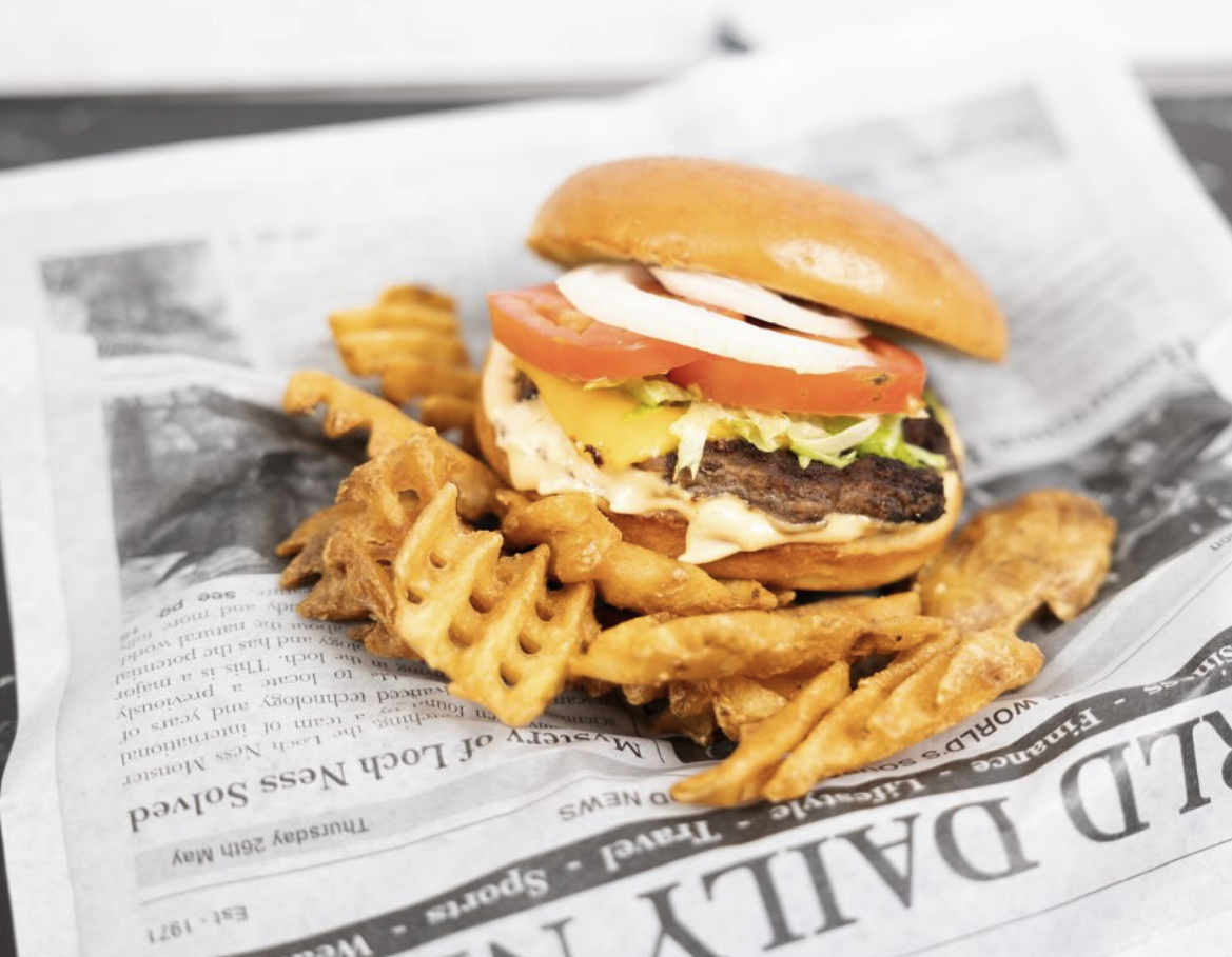 Hamburger with lettuce, tomato, cheese, and onion on a bun, served with waffle fries on a sheet of newspaper.
