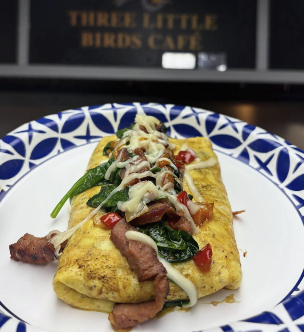 An omelette filled with spinach, bacon, chopped tomatoes, and melted cheese, served on a decorative blue and white plate, with a sign for 'Three Little Birds Cafe' in the background.