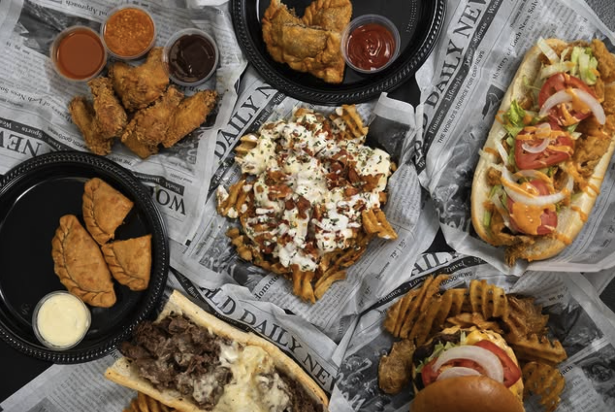 Assorted fast food including fried chicken tenders with dipping sauces, loaded fries with bacon and cheese, a hot dog with toppings, fried empanadas with dipping sauce, a cheeseburger, and a garlic parmesan chicken sandwich, all on newspaper-lined tr