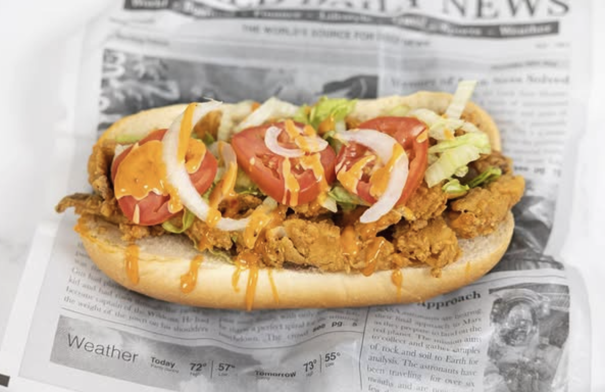 Fried chicken sandwich with lettuce, tomato, onions, and drizzle of sauce on a sandwich bun, placed on newspaper.