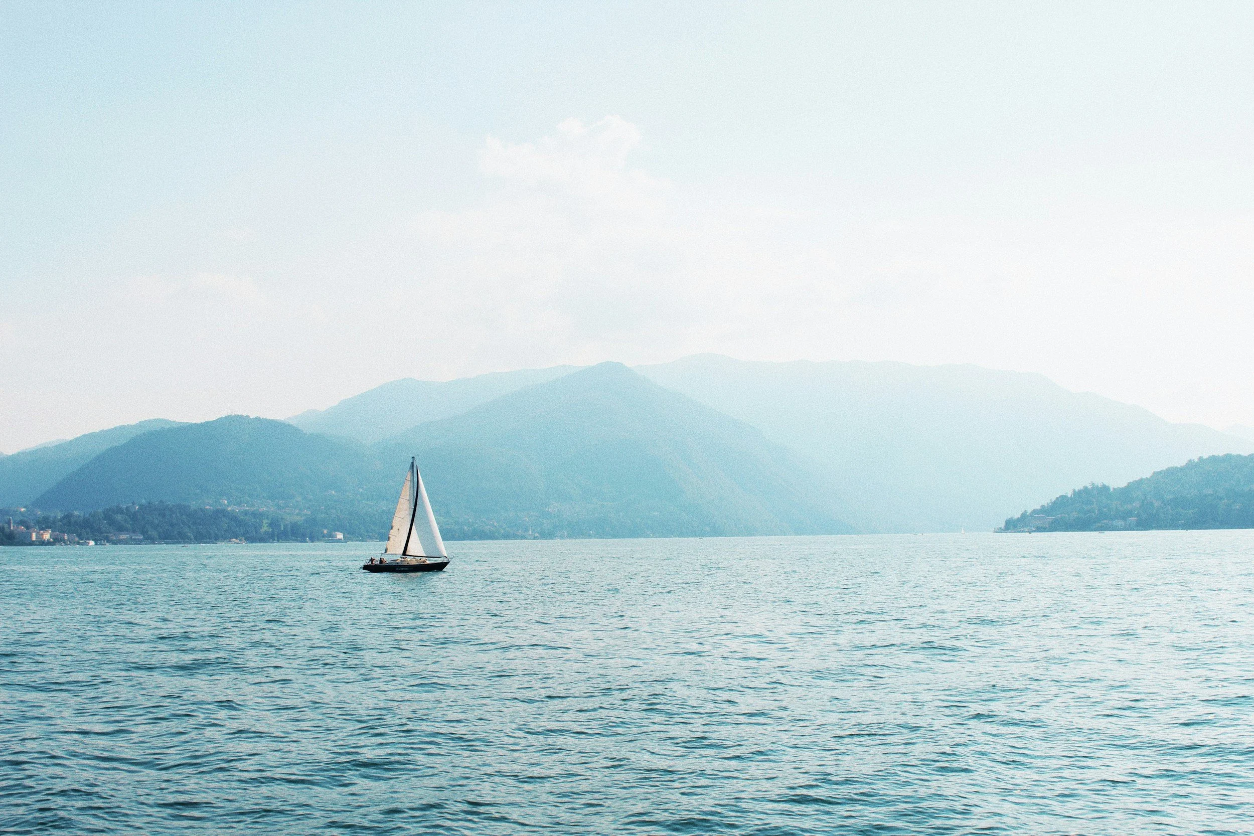 A sailboat on a calm body of water with distant mountains and a partly cloudy sky in the background.