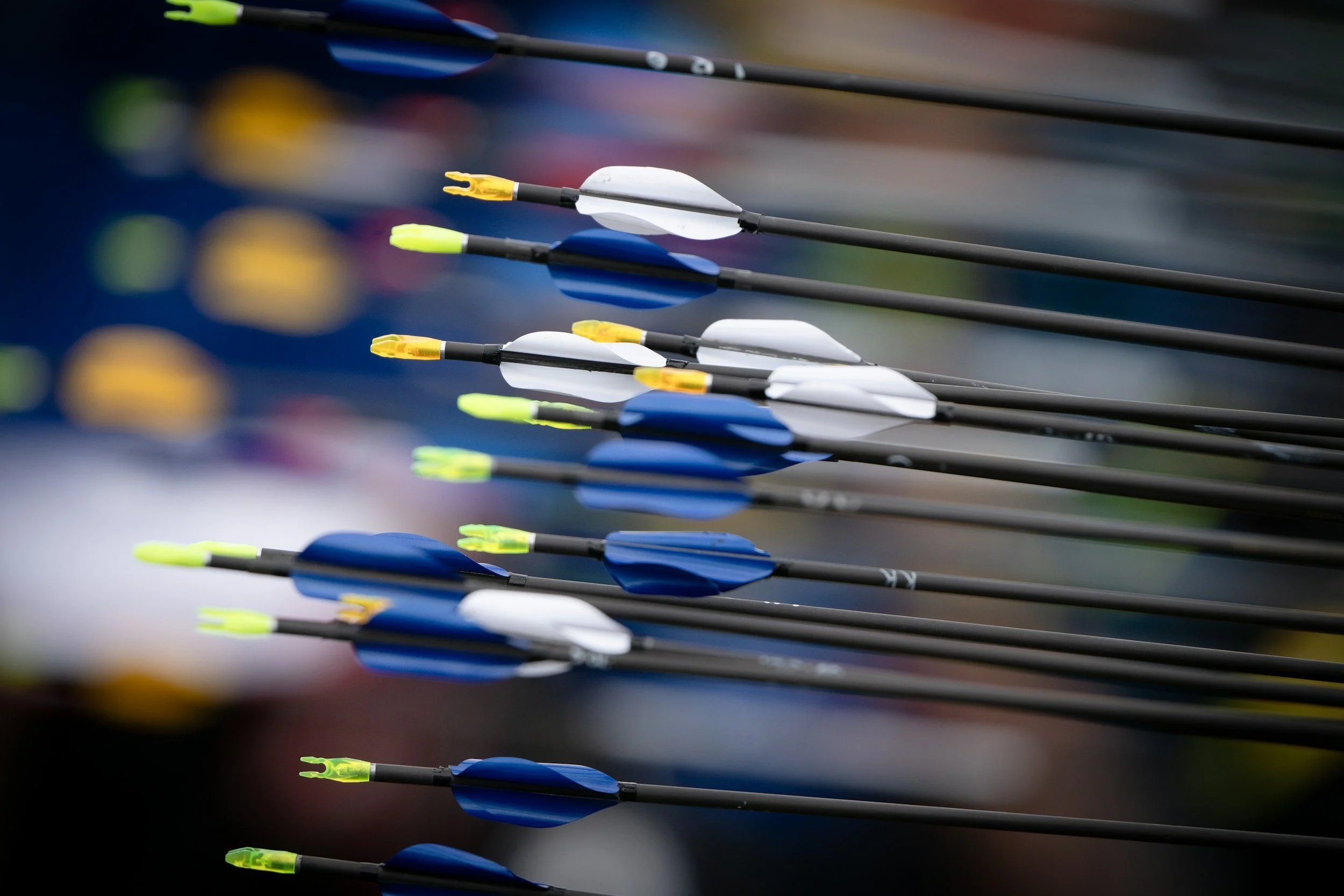 Close-up shot of multiple archery arrows with colorful fletching, lying parallel on a dark surface.