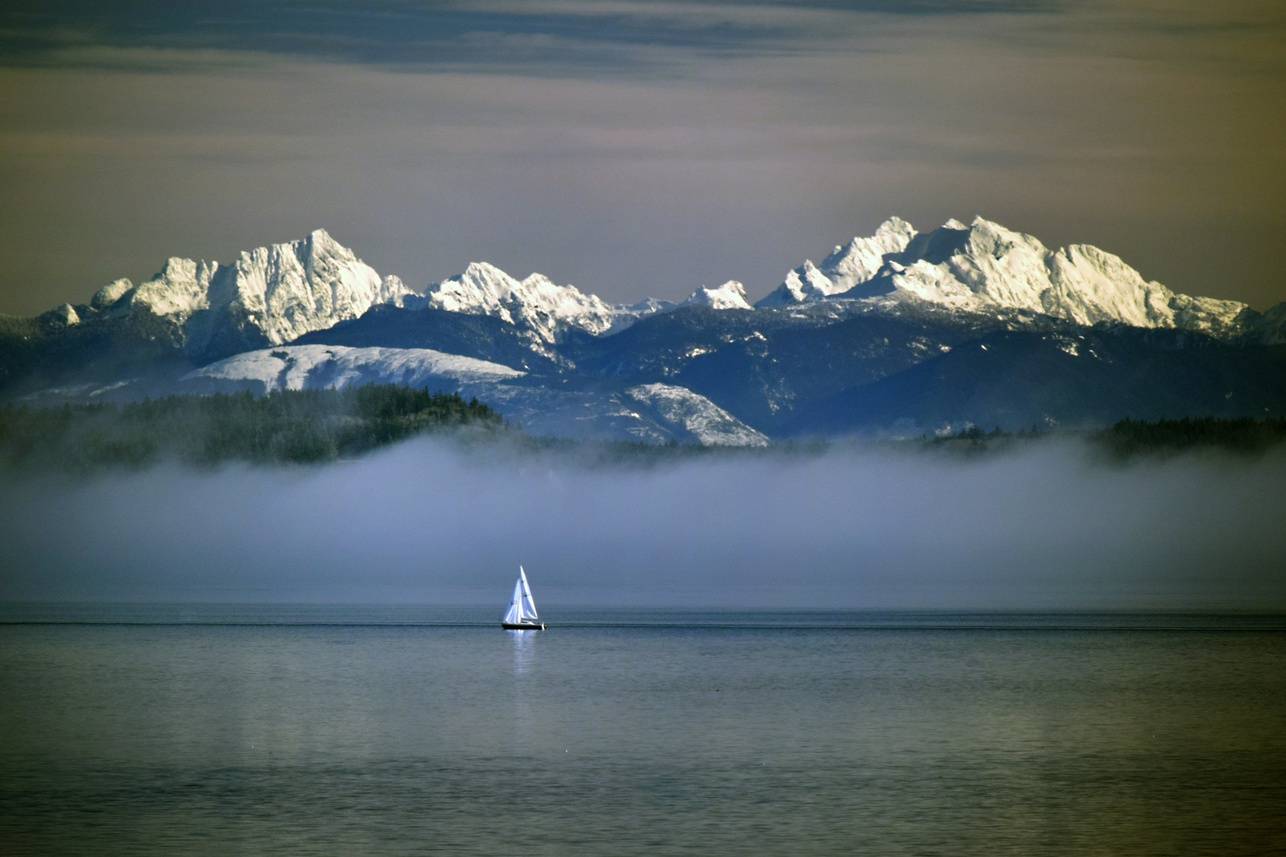 A sailboat with white sails sailing on a calm body of water, with snow-capped mountains in the background under a cloudy sky.