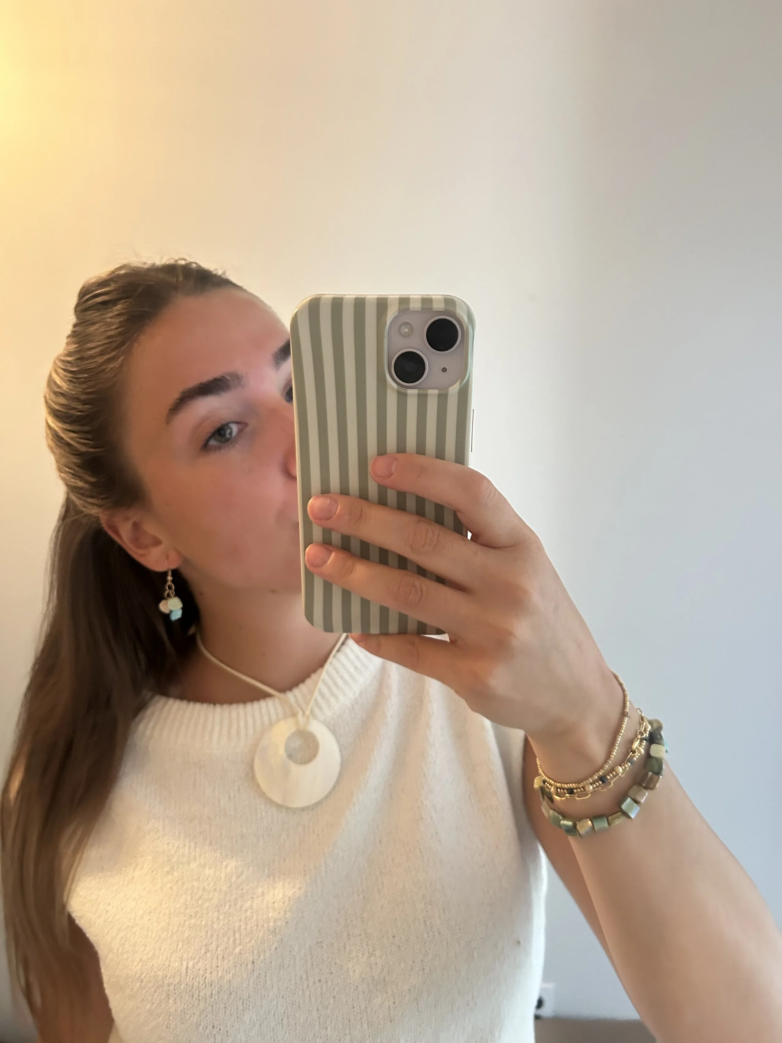 A woman taking a mirror selfie with a smartphone. Part of her face, shoulder, and arm are visible, she has long brown hair, wears earrings, a layered bracelet, a necklace with a large circular pendant, and a cream-colored knit top. The background is 