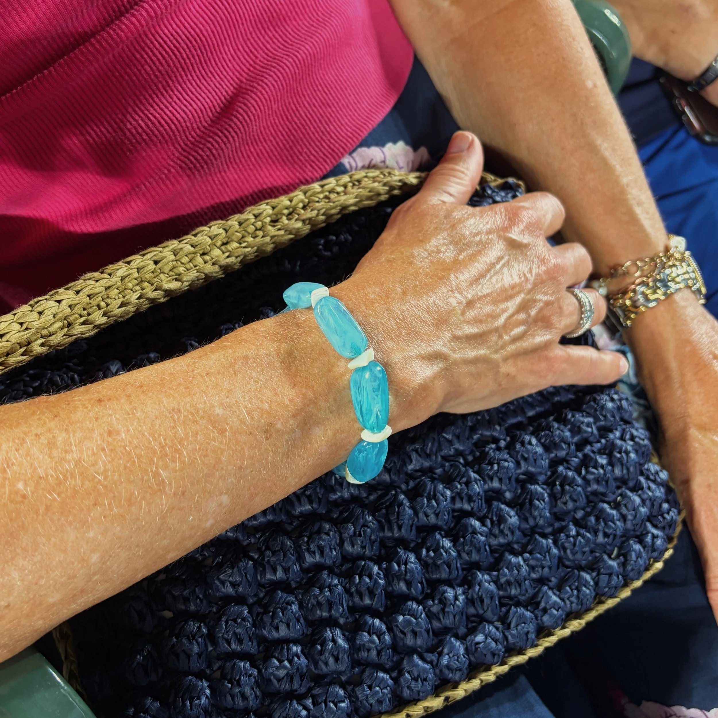 Close-up of a person's hand with jewelry and a blue bracelet, resting on a textured woven surface. The background includes a woman wearing a pink shirt and another with a green bracelet, along with a black textured fabric.