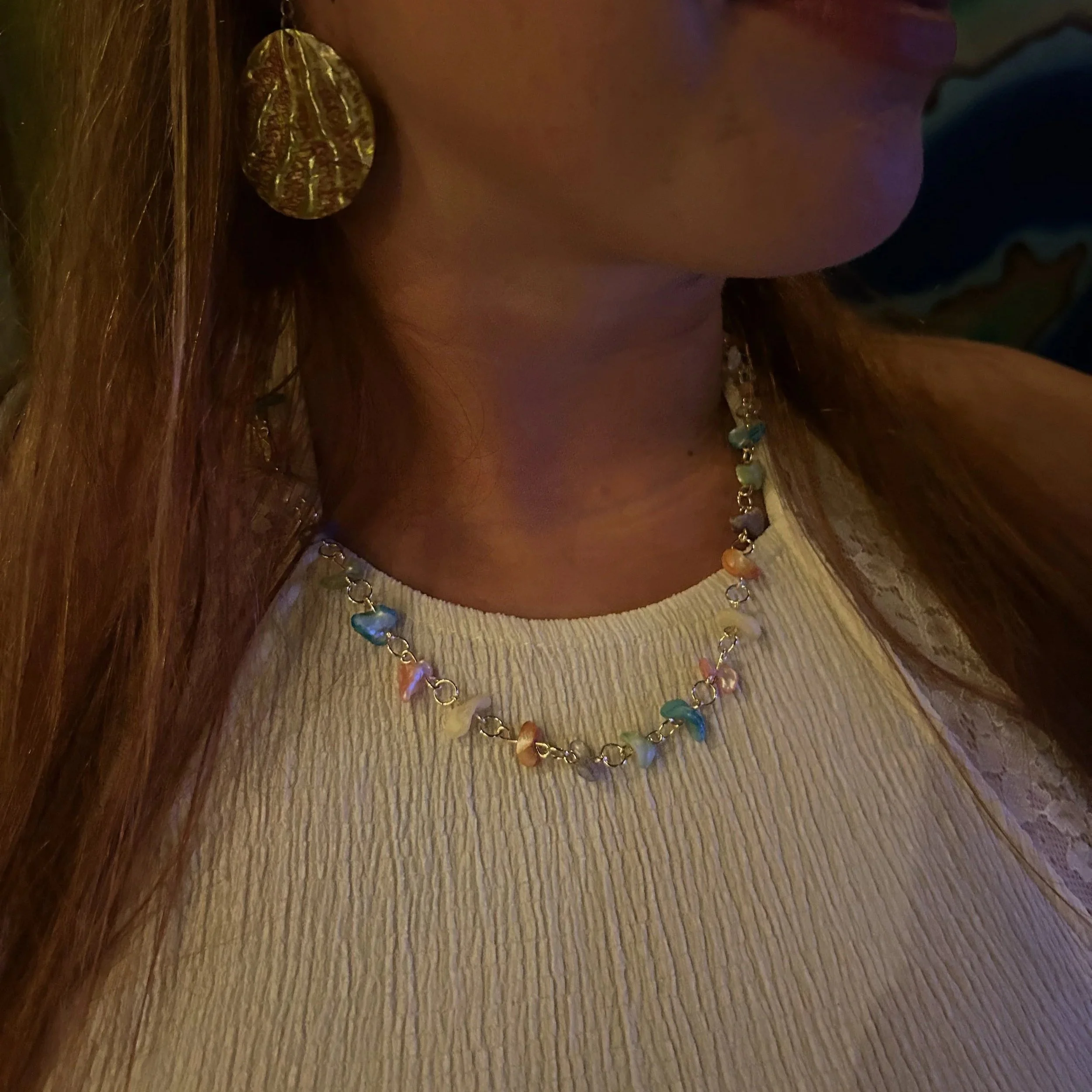 Close-up of a woman wearing gold earrings and a rainbow-colored beaded necklace, with long brown hair, standing against a plain background.