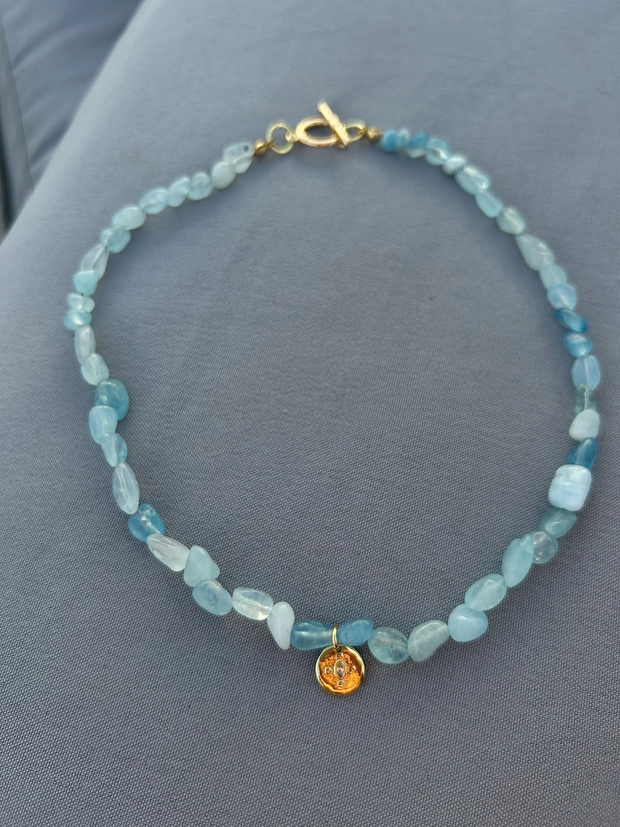 Blue and white beaded necklace with a gold circular pendant hanging from it, placed on a gray fabric surface.