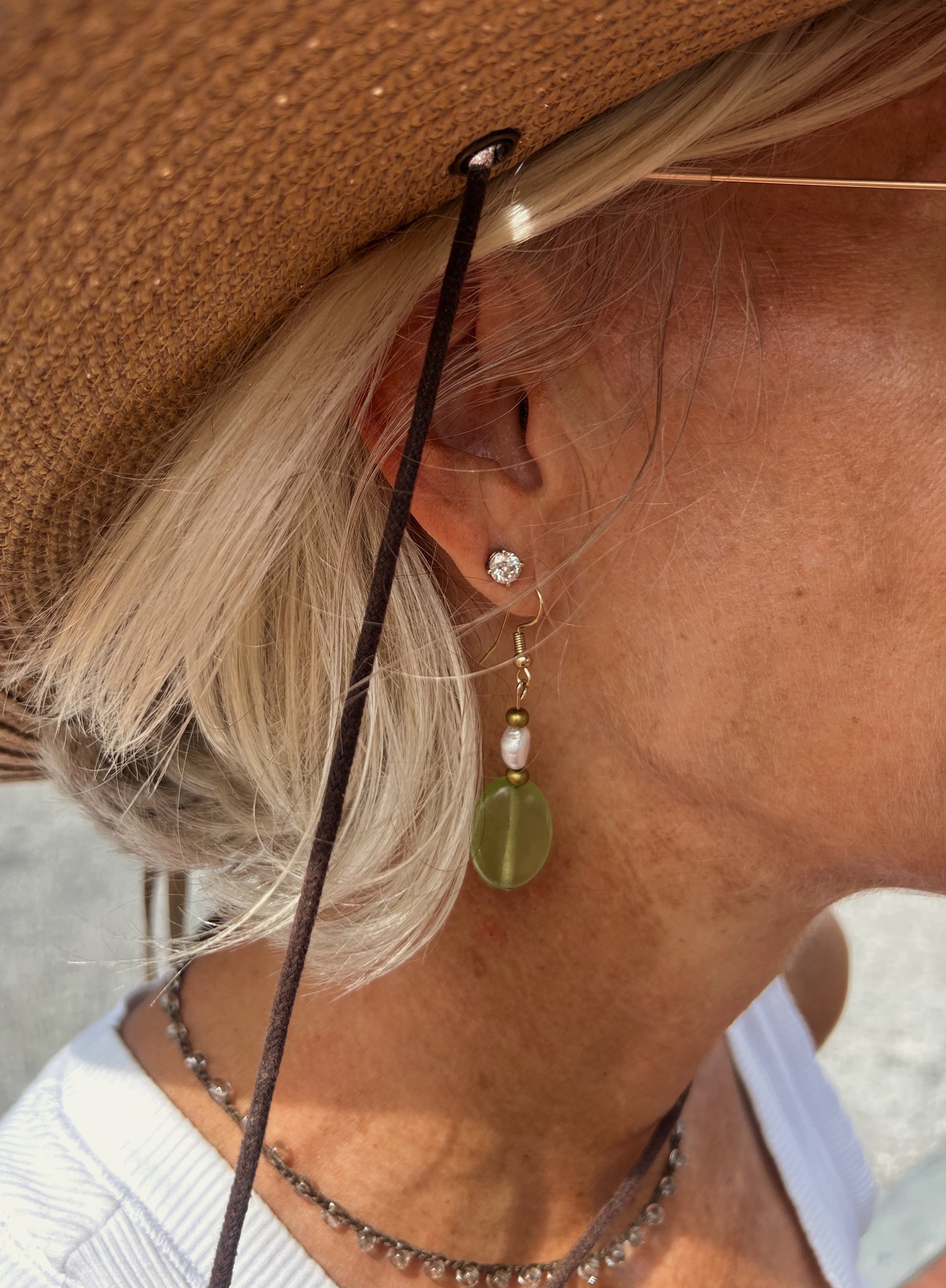 Close-up of a woman wearing a brown hat, earrings, necklaces, and a white top, with blonde hair and visible skin texture.