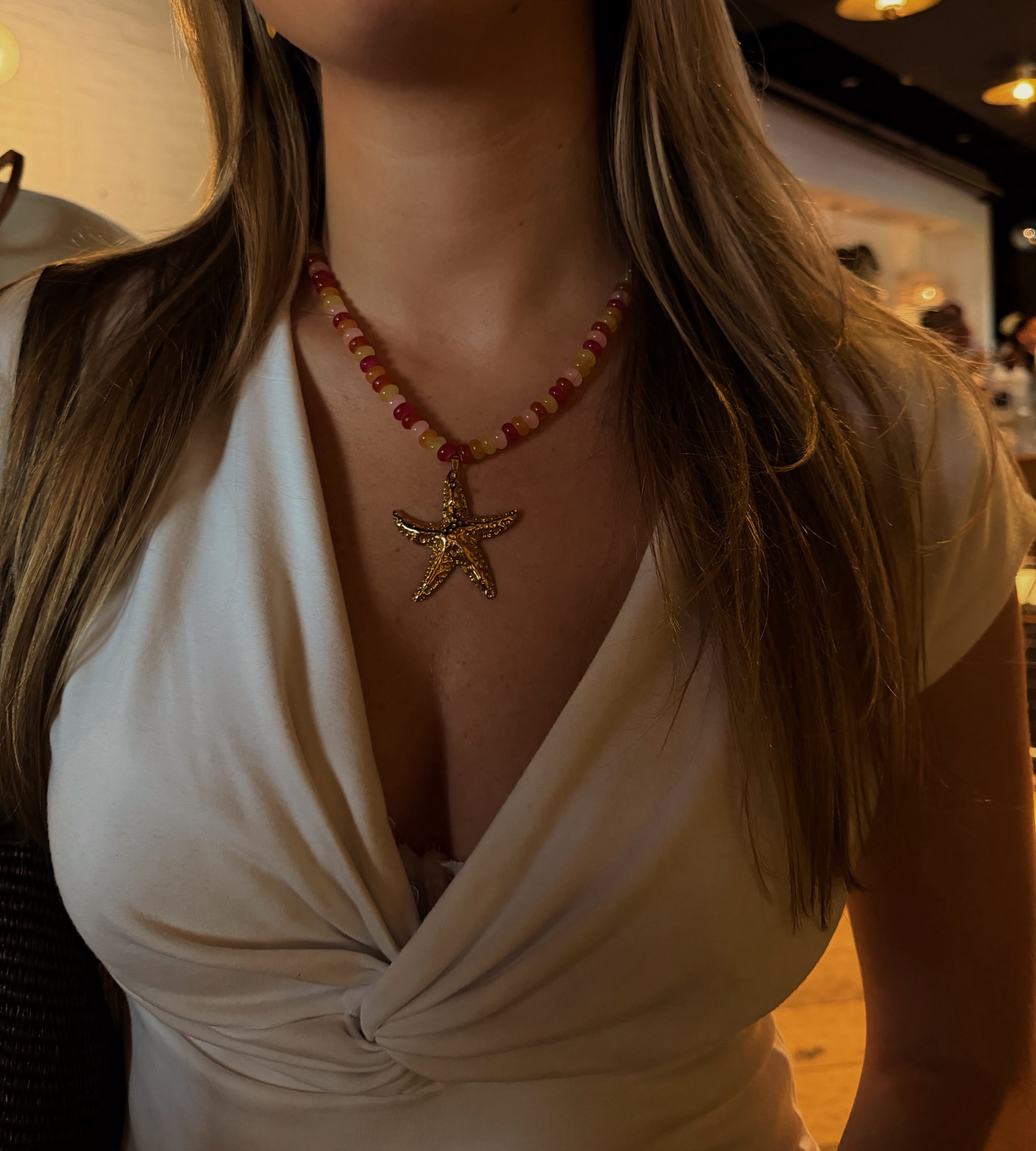 Close-up of a woman's upper body wearing a white dress with a deep neckline, a colorful beaded necklace with a star-shaped pendant, and long, wavy brown hair, in a dimly lit indoor setting.