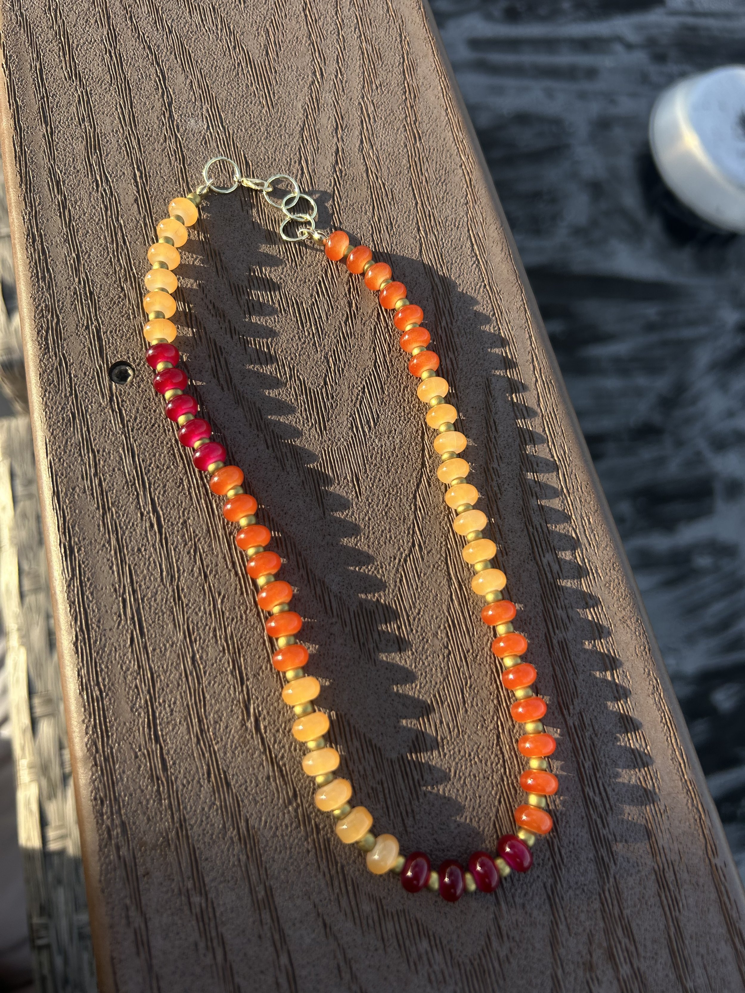 Colorful beaded necklace with shades of red, orange, yellow, and pink resting on a wooden surface near water.