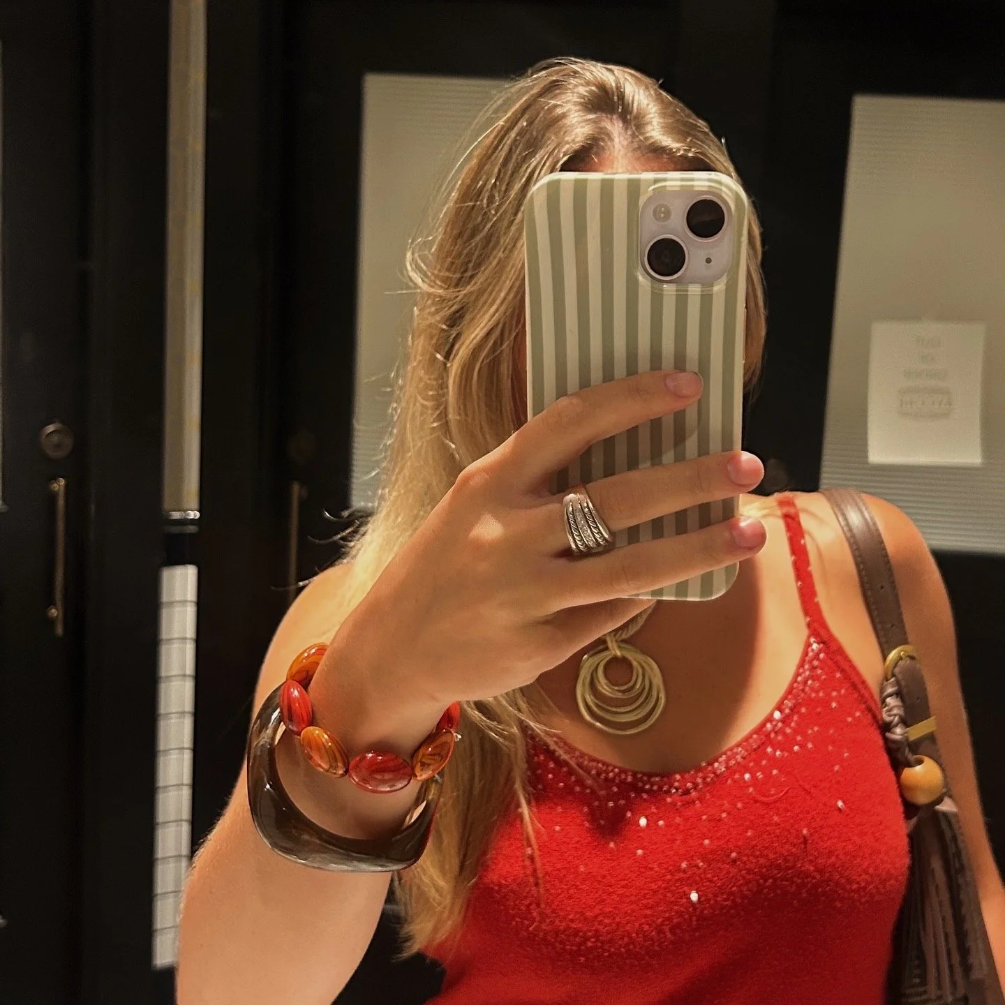 Person taking a mirror selfie with a smartphone, wearing a red top and jewelry, with long blonde hair, in a dark room.