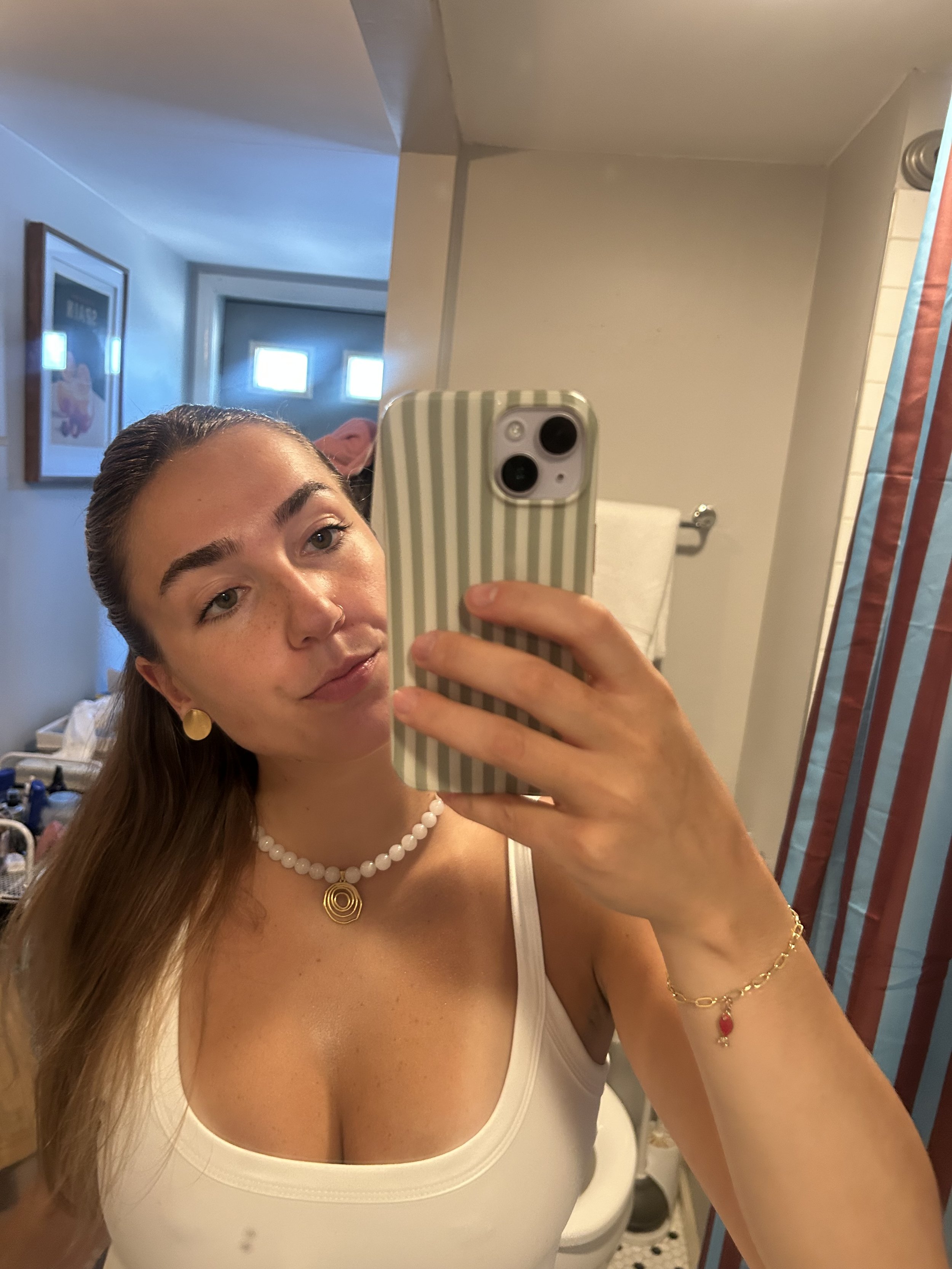 A young woman taking a mirror selfie in a bathroom, wearing a white tank top, pearl necklace, and gold jewelry, with her long hair down.