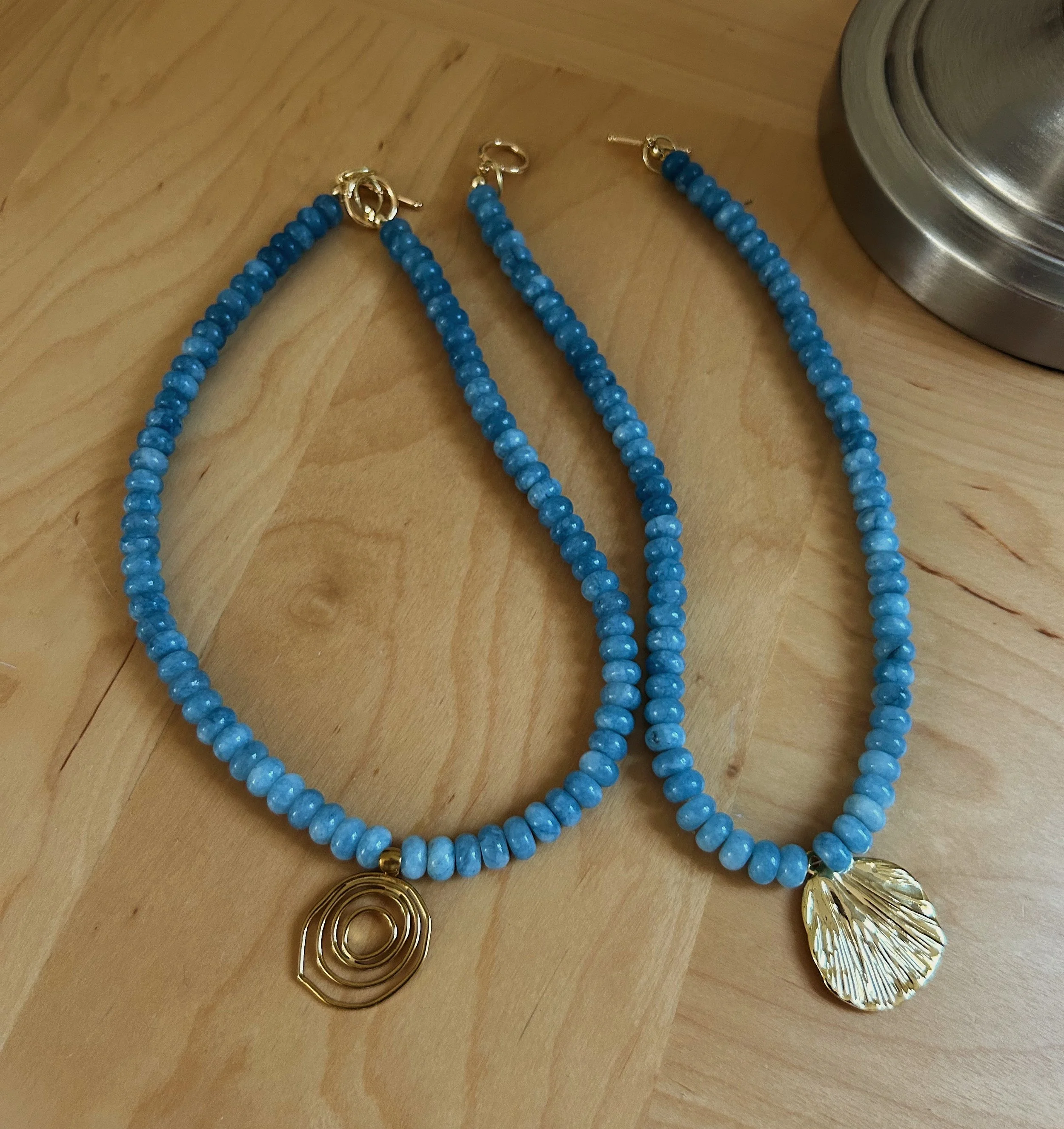 Blue beaded jewelry including earrings, rings, and a large gold-colored leaf-shaped pendant on a wooden surface.
