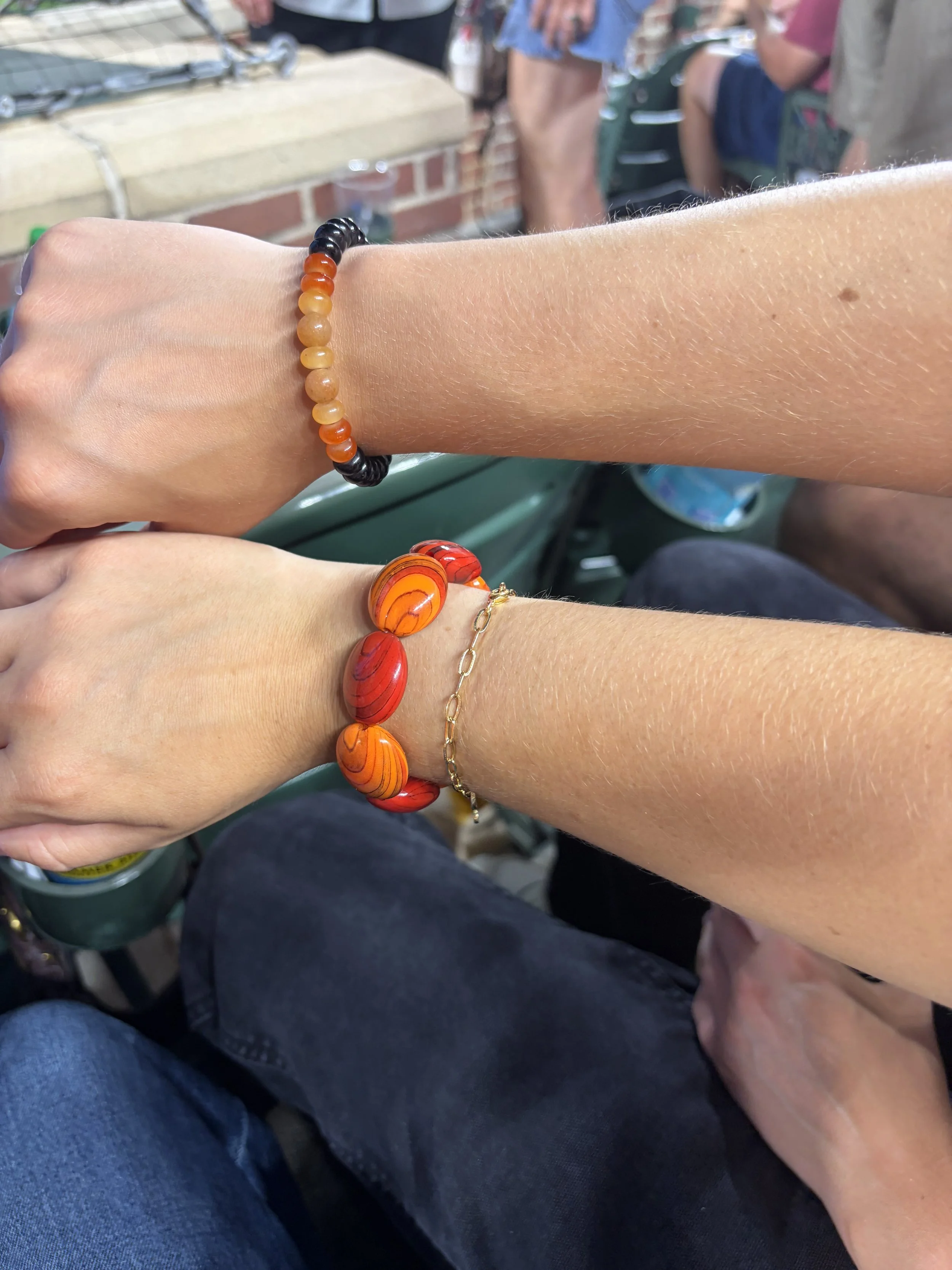 Close-up of two wrists with different bracelets, one with black and amber-colored beads, and the other with orange, red, and yellow beads, in an outdoor setting with people in the background.