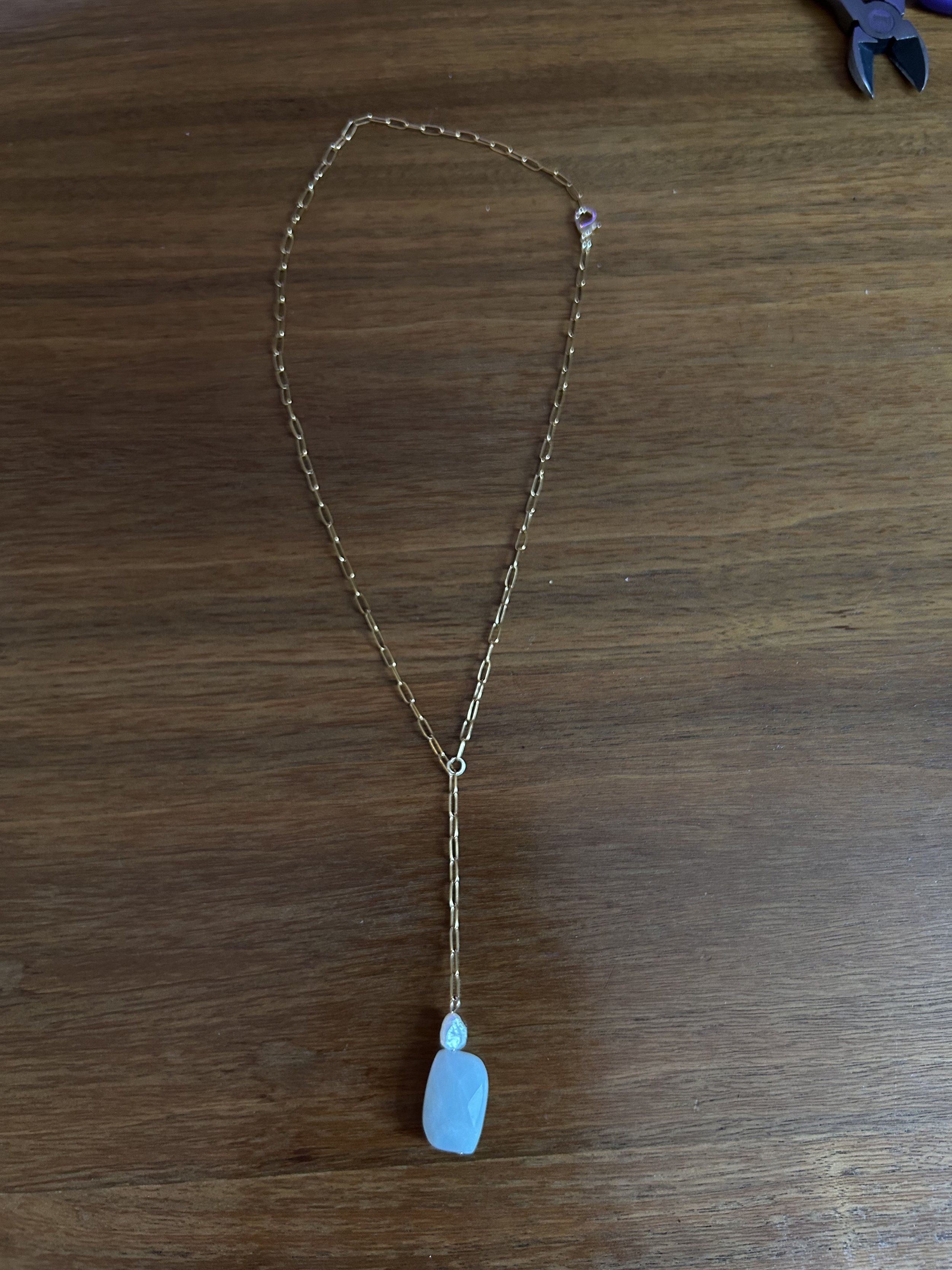 A gold chain necklace with a white pendant and a smaller blue flower-shaped detail, resting on a wooden surface.