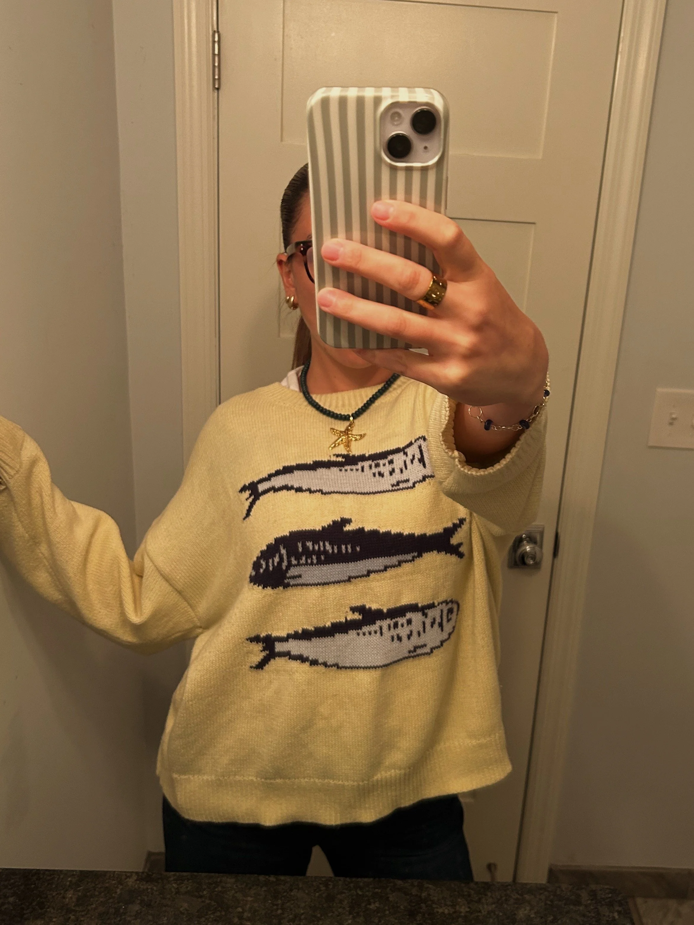 A person taking a mirror selfie in a bathroom, wearing glasses, a yellow sweater with a graphic design, and jewelry including a ring, necklace, and bracelet.