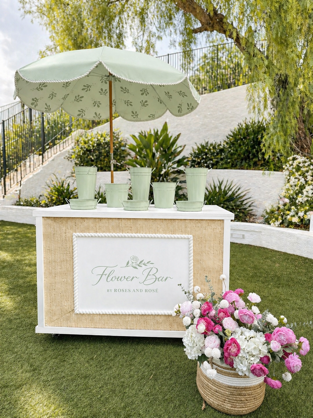 A flower bar stand with green watering cans and a green umbrella outdoors, decorated with pink and white roses and other flowers, in a garden setting.