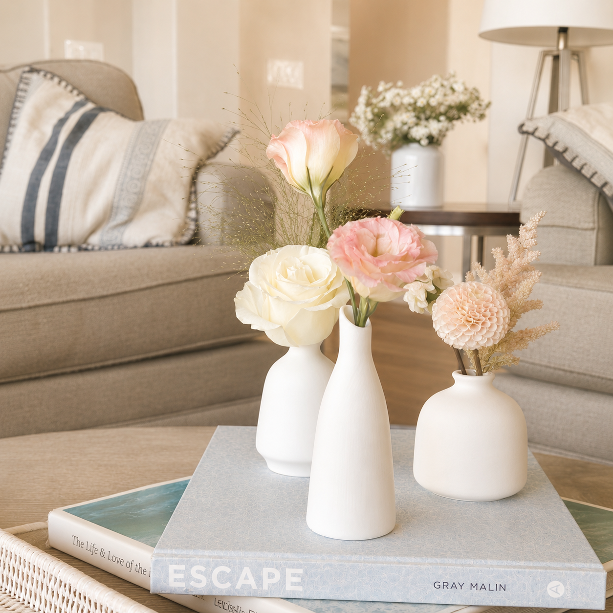 Three white vases with pink and white flowers on a light blue book titled 'ESCAPE' on a table in a living room with beige sofas and a pillow with a striped pattern.