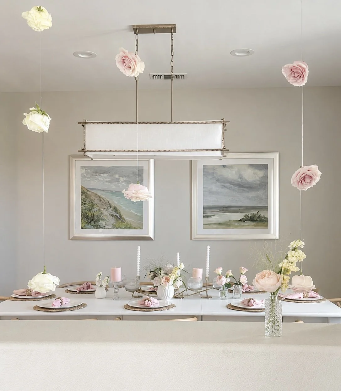 Decorated dining table with pink and white flowers, candles, and place settings, in front of two landscape paintings. Hanging floral accents from the ceiling.